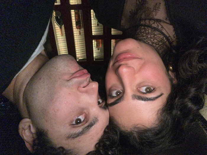 Two people pose closely together, their heads touching. The person on the left has short, dark hair, light skin, and light brown eyes. The person on the right has dark, curly hair, light skin, prominent eyebrows, and light brown eyes. They are both looking at the camera. The background features a wood-framed structure with shelves, possibly holding bottles or decorative items. The overall image has a slightly dark and warm tone. The couple is wearing dark clothing.