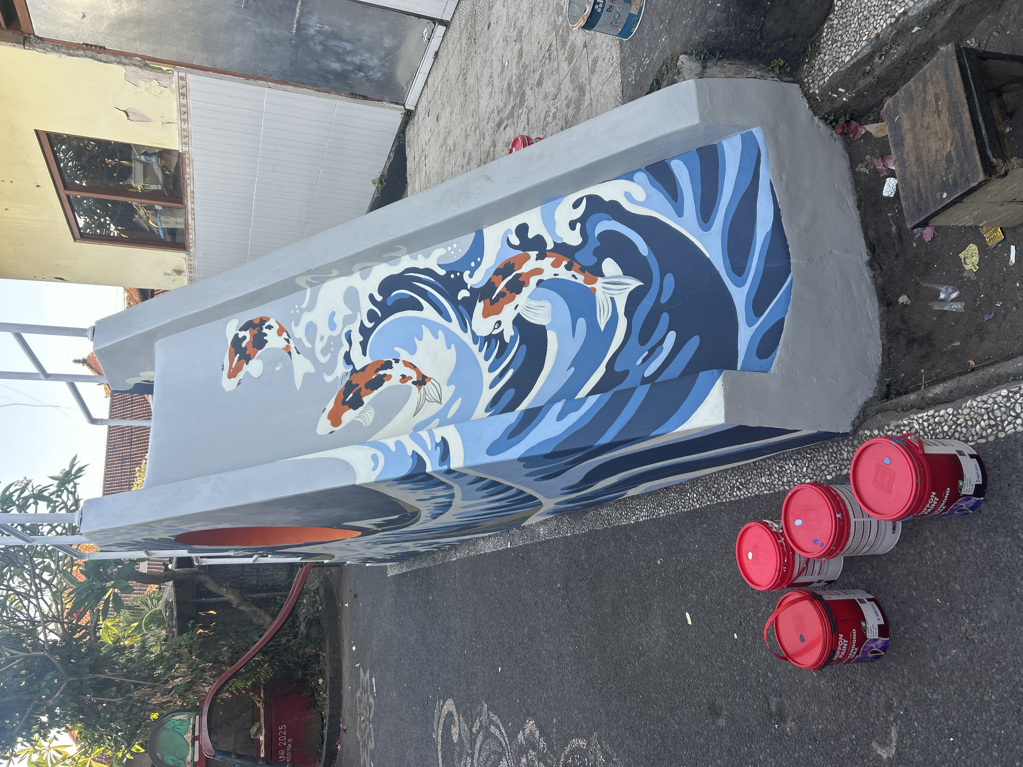The image depicts an outdoor scene where a grey playground slide has been painted with a mural. The mural features stylized blue waves and three koi fish in orange, white, and black. To the left of the slide, there's a red metal structure that appears to be part of an older playground. Four red paint buckets are on the asphalt ground in front of the slide. The slide is situated next to a building with a yellow facade, a brown window, and tiled lower walls. The ground is a mixture of asphalt and gravel. The environment appears to be a somewhat weathered urban or residential area.