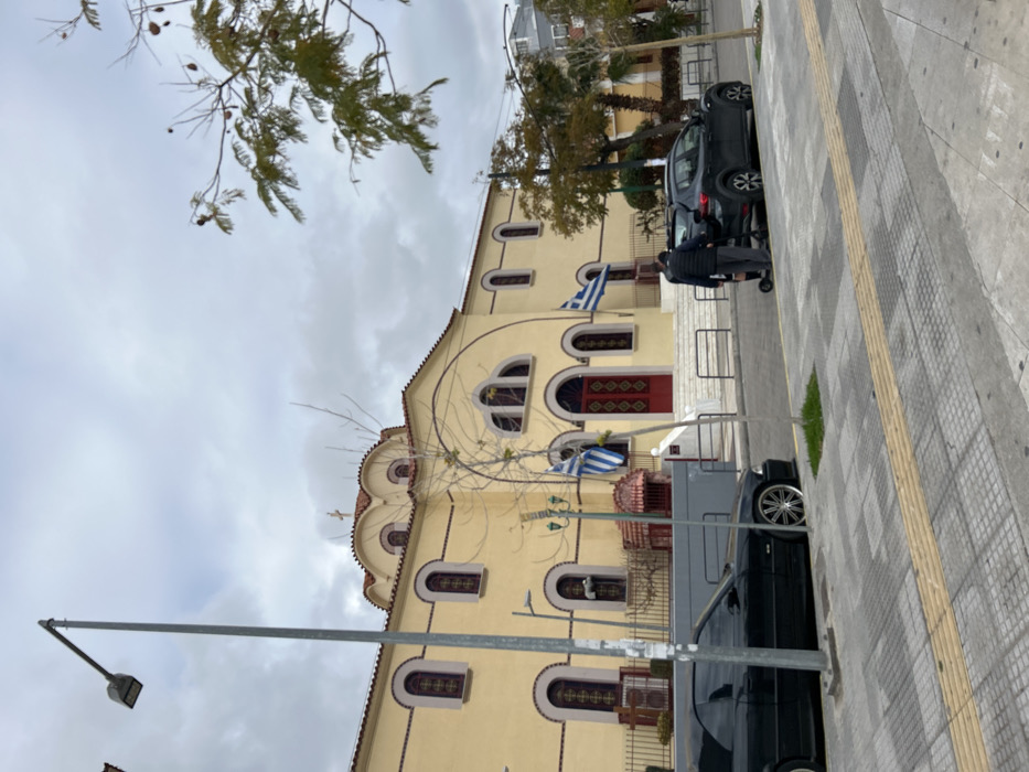 The image captures an outdoor scene in Nikea, Greece, centered on a large, light yellow Greek Orthodox church under an overcast sky. The church building features a red tiled roof with a visible cross atop one of its small domes or cupolas. Its facade is characterized by numerous arched windows, many appearing to have intricate decorative patterns or grilles, and a prominent red double door serving as the main entrance. Two Greek flags are proudly displayed on poles attached to the church wall, one above the entrance and another further to the left. Bare tree branches, possibly indicating late autumn or winter, extend into the upper and lower left portions of the frame, partially obscuring the church.
In the foreground and to the right, a paved sidewalk with alternating grey and tan squares, along with a yellow tactile strip, runs parallel to a street. A black SUV is parked on the street next to the sidewalk. A person, dressed in dark attire, stands near the rear of the SUV, appearing to interact with its trunk or cargo area. Another dark-colored vehicle is partially visible behind a grey metal street light pole in the lower left. The sky is uniformly cloudy, suggesting a daytime scene with diffused light. No discernible text is visible within the image.