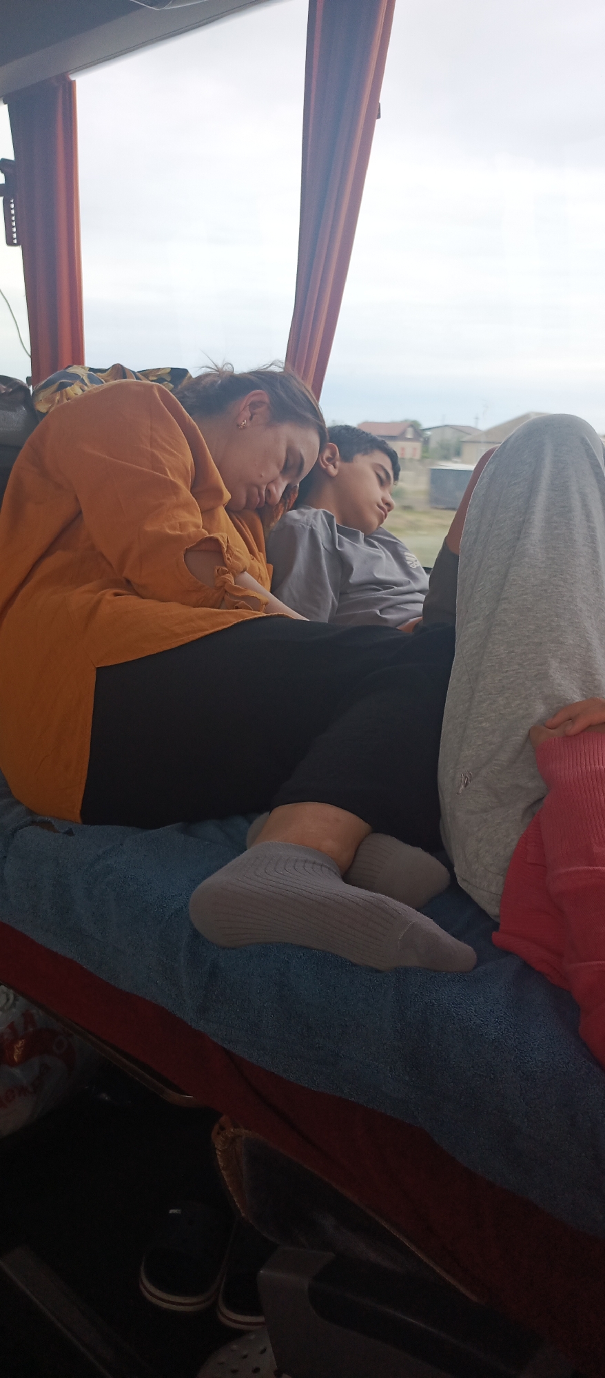 In this indoor scene, two individuals, likely a mother and son, are asleep on a bus or train. The woman, on the left, wears an orange blouse and dark pants, her head resting on her arm. Beside her, a boy in a grey t-shirt also sleeps, his head leaning slightly to the right. The foreground shows their legs covered in socks and a blanket. The background reveals a window with a view of a suburban landscape with houses and vegetation under an overcast sky. The seating appears to be upholstered in blue fabric, and there's a red bag in the lower right corner of the frame.
