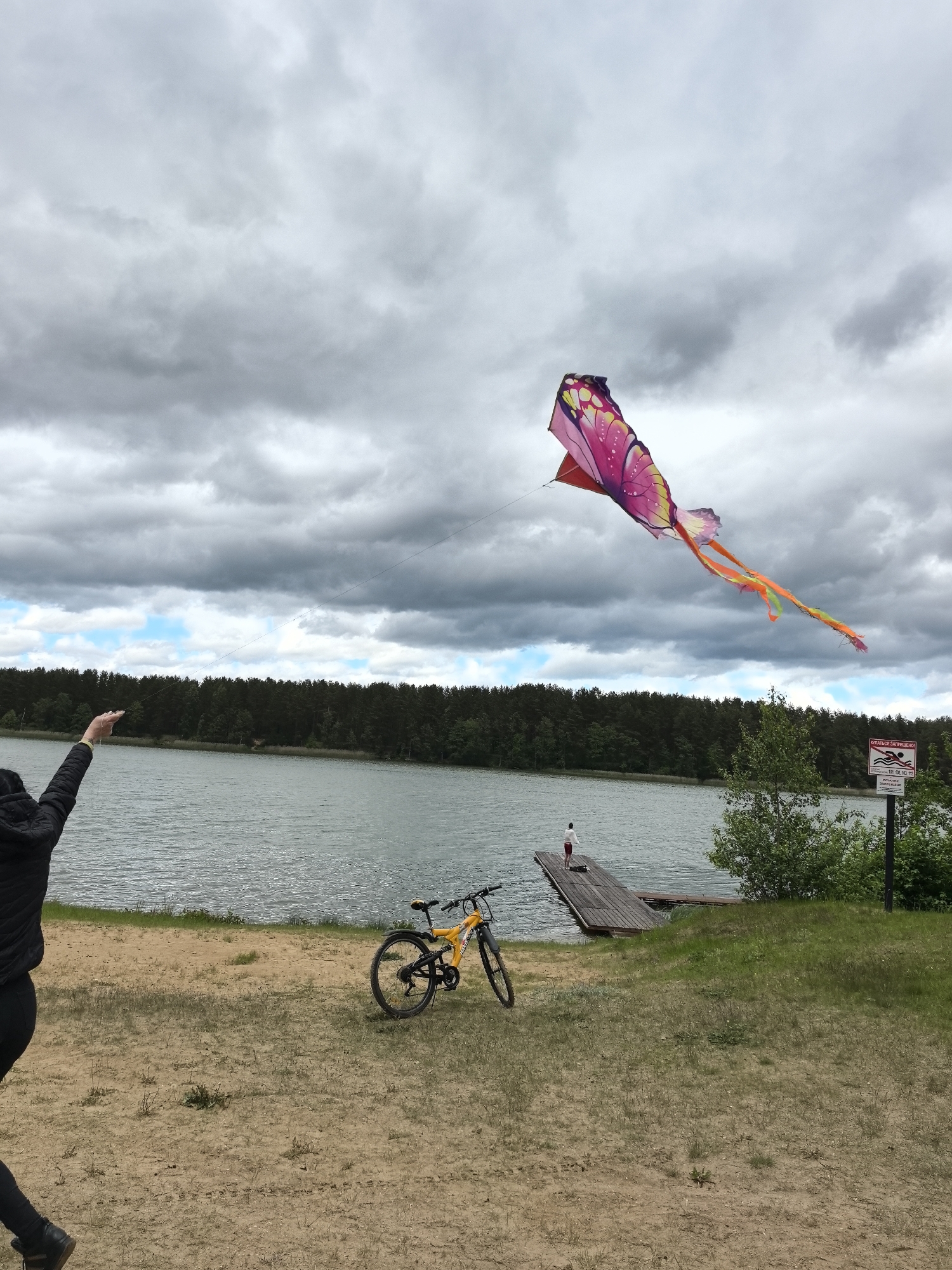 The image depicts a lakeside scene in Парэчча, Belarus, under a cloudy sky with patches of blue. A person stands on the left, partially visible, with their arm raised, presumably flying a large, butterfly-shaped kite which dominates the upper right portion of the frame. The kite is brightly colored with pink, red, and orange hues. In the background, a body of water stretches across the frame, bordered by a dense forest. A wooden dock extends into the water, with a person standing at the end of it. A yellow bicycle is propped on the grassy area near the beach. A sign, possibly indicating swimming regulations, is visible on the right side of the frame. The foreground consists of a sandy beach transitioning into a grassy area.