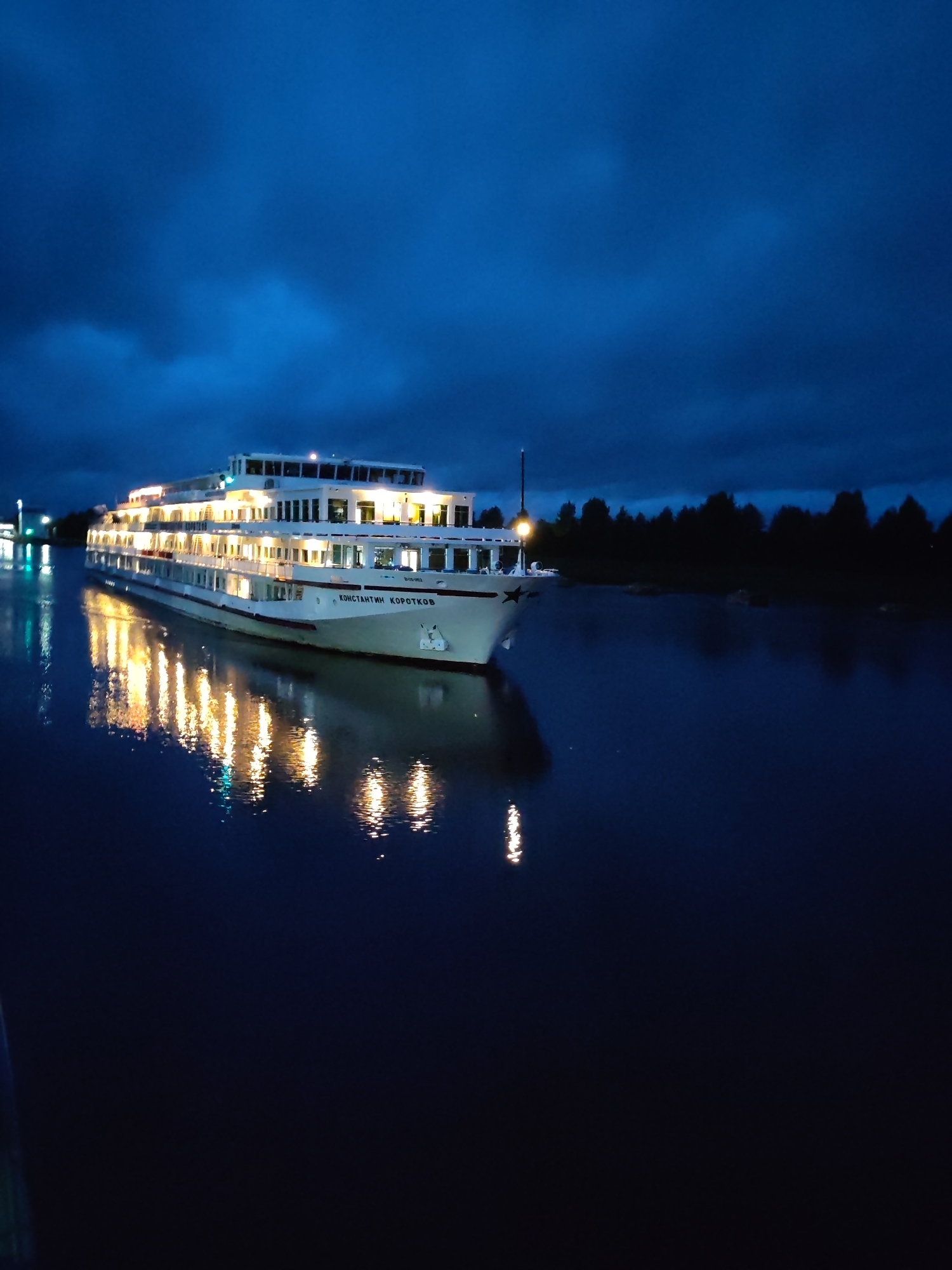 A large white river cruise ship named "KONSTANTIN KOROTKOV" is docked on a dark blue body of water, likely a river, under a deep blue, cloudy night sky. The ship is brightly illuminated from within, with numerous windows glowing warm yellow. Its reflection is clearly visible on the still water, creating a mirrored effect of the lights. The ship has multiple decks and a distinctive profile with a star emblem on its bow. A dark, silhouetted treeline forms the background on the right side of the image, suggesting a natural landscape. On the left side, a distant structure with lights is visible, possibly a dock or another building. The overall atmosphere is calm and serene, despite the dark sky. The location is identified as Devyatiny, Russia. There are no people visible in the image.