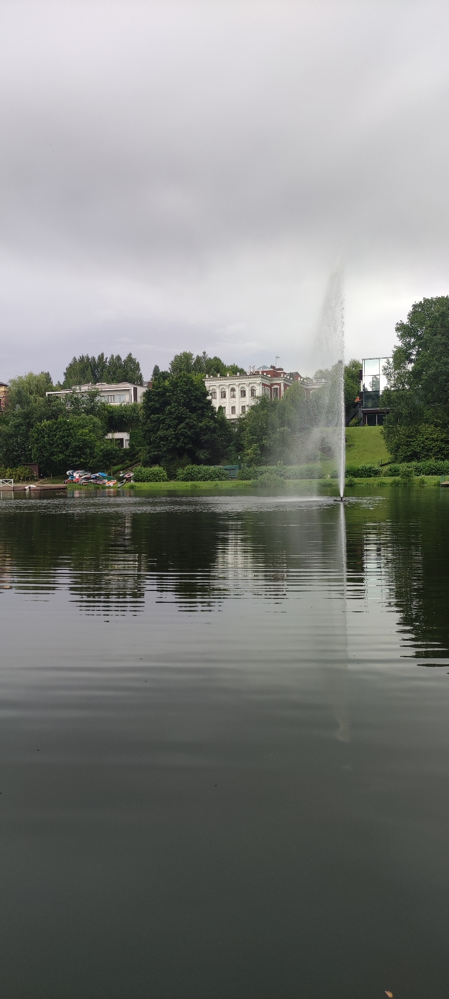 The image captures a tranquil scene of a lake with a tall fountain spewing water high into the air. The lake's surface is rippled, reflecting the overcast sky and the lush green trees that line the shore. Behind the trees, several multi-story buildings are visible, including a large white building with intricate architectural details and a more modern structure with glass walls. On the left side of the image, a small dock is visible, with several small boats and kayaks neatly arranged on it. The overall atmosphere is peaceful and serene, suggesting a well-maintained park or residential area. The location is specified as Angelovo, Russia.