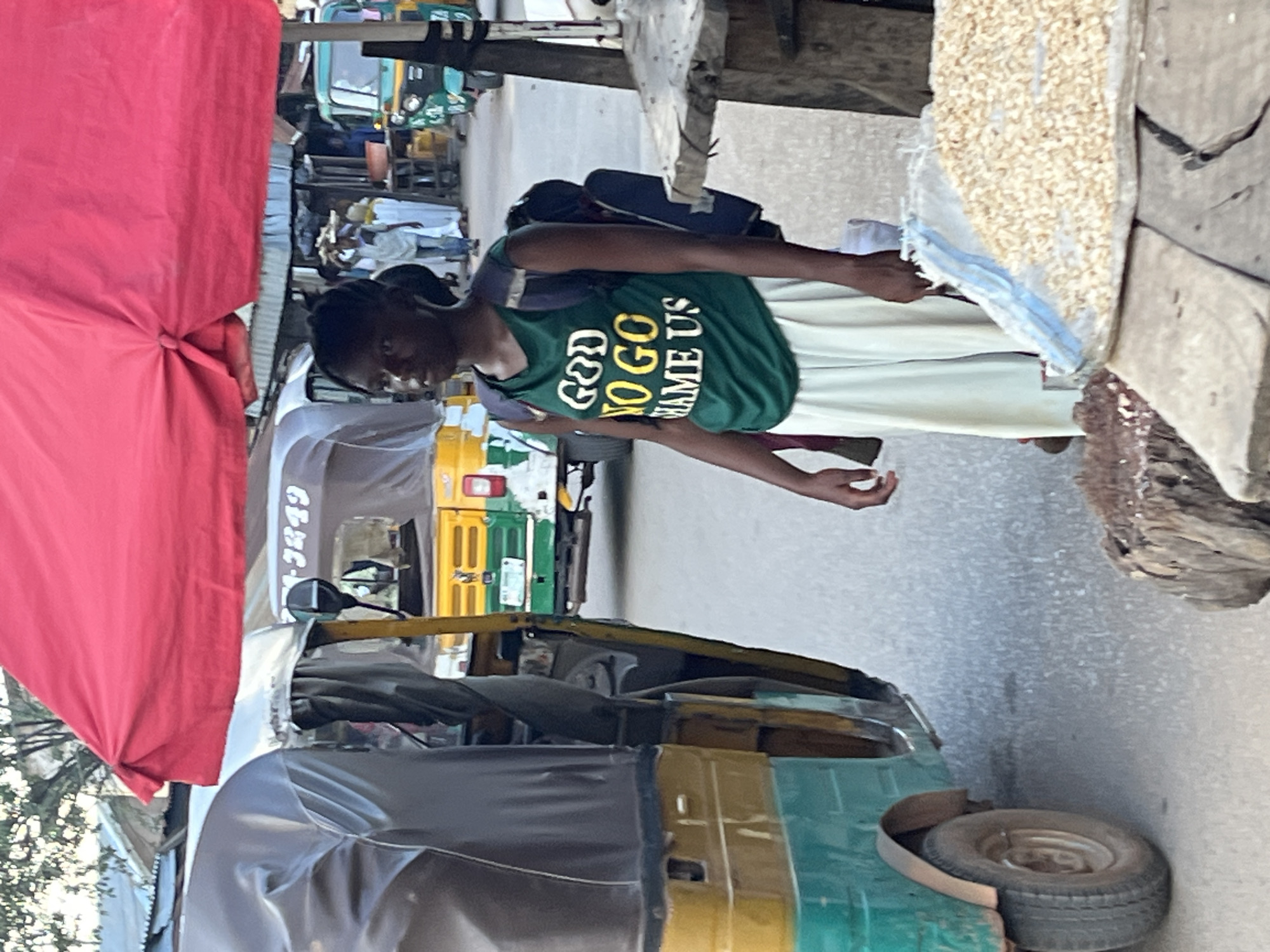 The image captures a street scene in Jos, Nigeria. A young person with braided hair is the central subject, wearing a green t-shirt with white and yellow text that reads "GOD NO GO SHAME US". They are also wearing a light-colored skirt and a backpack. The person appears to be standing and interacting with someone off-camera, with their hands outstretched.
In the background, several auto-rickshaws, a common mode of transportation in Nigeria, are visible. One is predominantly green and yellow, another is white, and a third is green and white. Some appear to be parked or waiting for passengers. A large red tarp covers a portion of the left side of the frame, possibly indicating a market stall or temporary shelter. A sack filled with light-colored grains is propped up on the right side of the image.
The setting suggests a busy outdoor environment, likely a market or a transportation hub. The lighting indicates it's daytime, though the specific time of day and weather conditions are not clearly discernible. The overall mood appears to be casual and everyday.