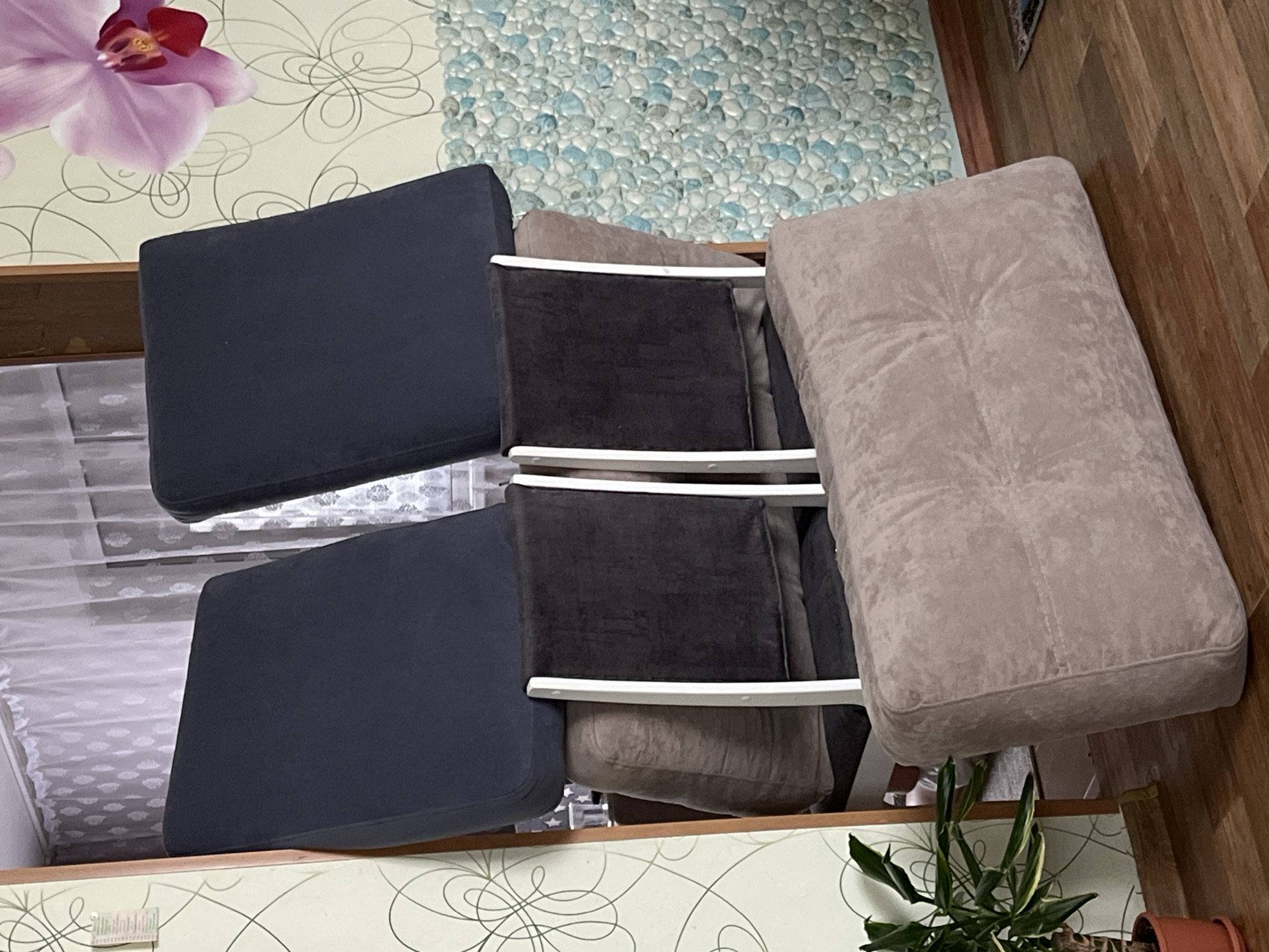 The image shows three chair cushions placed on top of each other. The top cushion is light brown and rectangular. The middle cushion is dark brown. The bottom cushion is navy blue. The chair frames are white. The cushions are stacked on a surface with a floral and swirl design. The background includes a wood floor, a wall with a pebble-like design, and a bed. A potted plant is also visible. It is impossible to determine the specific location within Сосново, Russia from the given image.