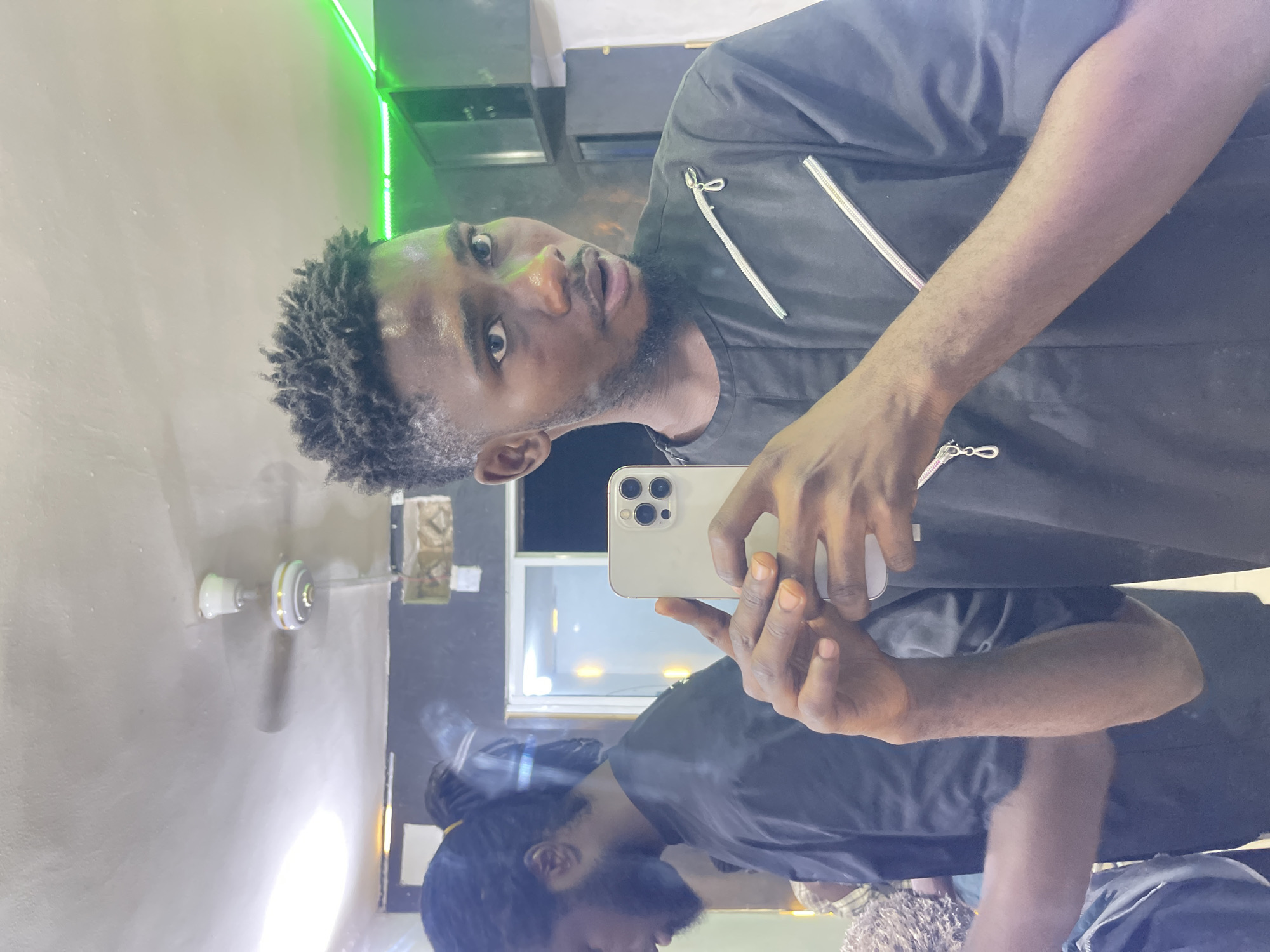 The image displays a mirror selfie of a dark-skinned man captured indoors. The photograph is oriented horizontally, showing the man looking upwards and slightly to the right from the left side of the frame. He has short, textured dark hair and a beard. He is wearing a dark, possibly black or charcoal, short-sleeved shirt featuring prominent silver zippers on the chest.
The man holds a light-colored smartphone, likely an iPhone due to its triple camera array, in both hands, positioned to capture his reflection. His reflection is visible below the phone and his hands, showing the top of his head, part of his face, and his shirt.
The background of the mirror selfie reveals elements of an indoor space. A light-colored ceiling with a white and gold ceiling fan is visible on the far left. Towards the upper right, a dark wall is illuminated by bright green LED strip lights running vertically. In the reflection, a window or door frame with a white border can be seen, indicating another light source or room beyond. The overall lighting is a mix of artificial illumination from the green lights and ambient light reflected in the mirror. The man's expression is neutral, as he looks directly into the phone's camera. No discernible text is visible in the image.