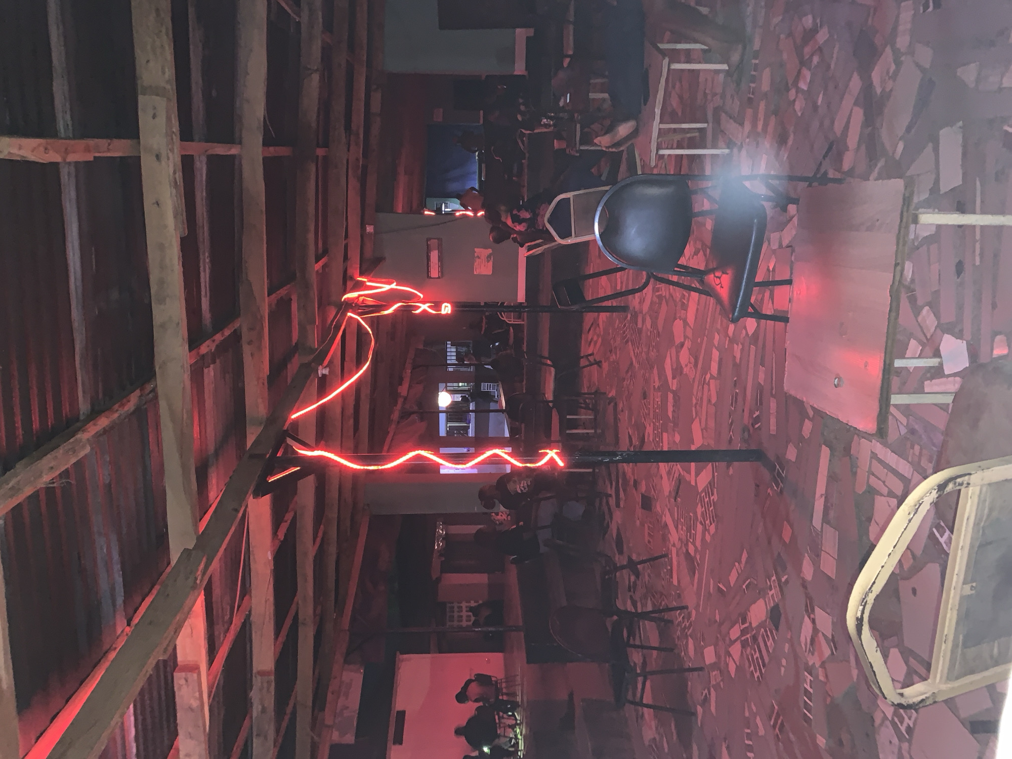 The image depicts an interior scene, possibly a bar or restaurant, illuminated by red neon lights strung across a wooden beam ceiling. The location appears to be in Kabong, Nigeria, based on provided context. Patrons are seated at tables, with chairs of various designs. The floor is tiled with a geometric pattern, predominantly in red hues. Shadows are prominent, obscuring details of the individuals and their interactions. Overall, the atmosphere appears dimly lit and social.