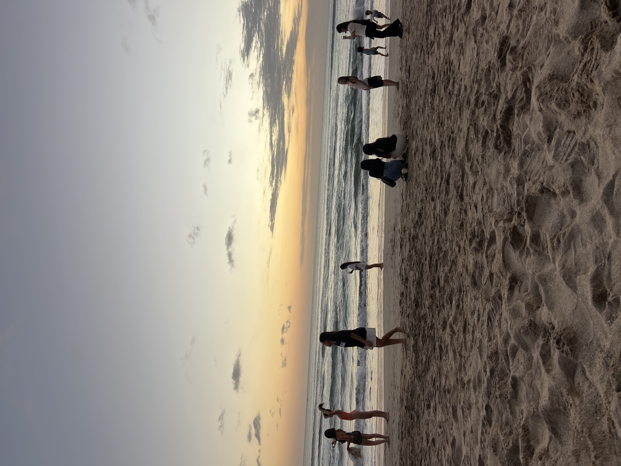 The image shows a beach scene at sunset in Kuta, Indonesia. The sky is a gradient of pale blue and orange, with scattered clouds. The ocean has moderate waves, and the beach is sandy with some textured patterns in the foreground. There are several people on the beach, appearing to be tourists enjoying the sunset.
In the foreground, the sand is the dominant element. In the mid-ground, several groups and individuals are visible. On the left, a woman in a bikini and another in a coral-colored swimsuit are standing. Further right, a woman in a black t-shirt and white skirt is walking, and another woman is standing and looking out at the sea. Towards the right side of the image, a group of four individuals is seated on the sand, and a woman in a white top and dark pants is standing and holding a phone, possibly taking a photo. A child in a blue shirt is visible walking near the seated group. In the background, more people are scattered along the shoreline and in the water.
The overall mood is relaxed and serene, typical of a beach sunset. The lighting suggests it is late afternoon or early evening.