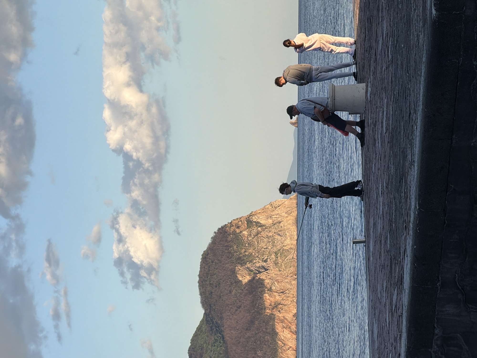 The image captures a coastal scene in Petrovac, Montenegro, at what appears to be late afternoon or early evening, characterized by warm light on the landscape. The photograph is taken from a vantage point on a concrete pier or seawall, looking out over the sea towards a prominent mountain.
Four individuals are depicted standing on the pier. On the left, one person, dressed in a light grey hoodie and dark pants, is actively engaged in fishing, holding a fishing rod that extends towards the water. Further along the pier, a second person, wearing a light shirt, shorts, and a cap, is leaning over the edge, looking down at the water. Behind them, a third individual in a grey jacket and pants is also observing the sea. The fourth person, positioned further to the right, is wearing light-colored clothing and also appears to be focused on the water. A white, cylindrical object is also visible on the pier between two of the individuals.
The body of water, likely the Adriatic Sea, exhibits a calm, slightly rippled surface. Across the water, a large, rocky mountain rises, its slopes covered in sparse, dry vegetation and exposed rock, bathed in the soft, warm glow of the setting sun. The sky above is a clear blue, adorned with scattered white and grey clouds, some illuminated by the same warm light. The scene conveys a tranquil atmosphere, highlighting leisure activity by the sea. There is no visible text in the image.