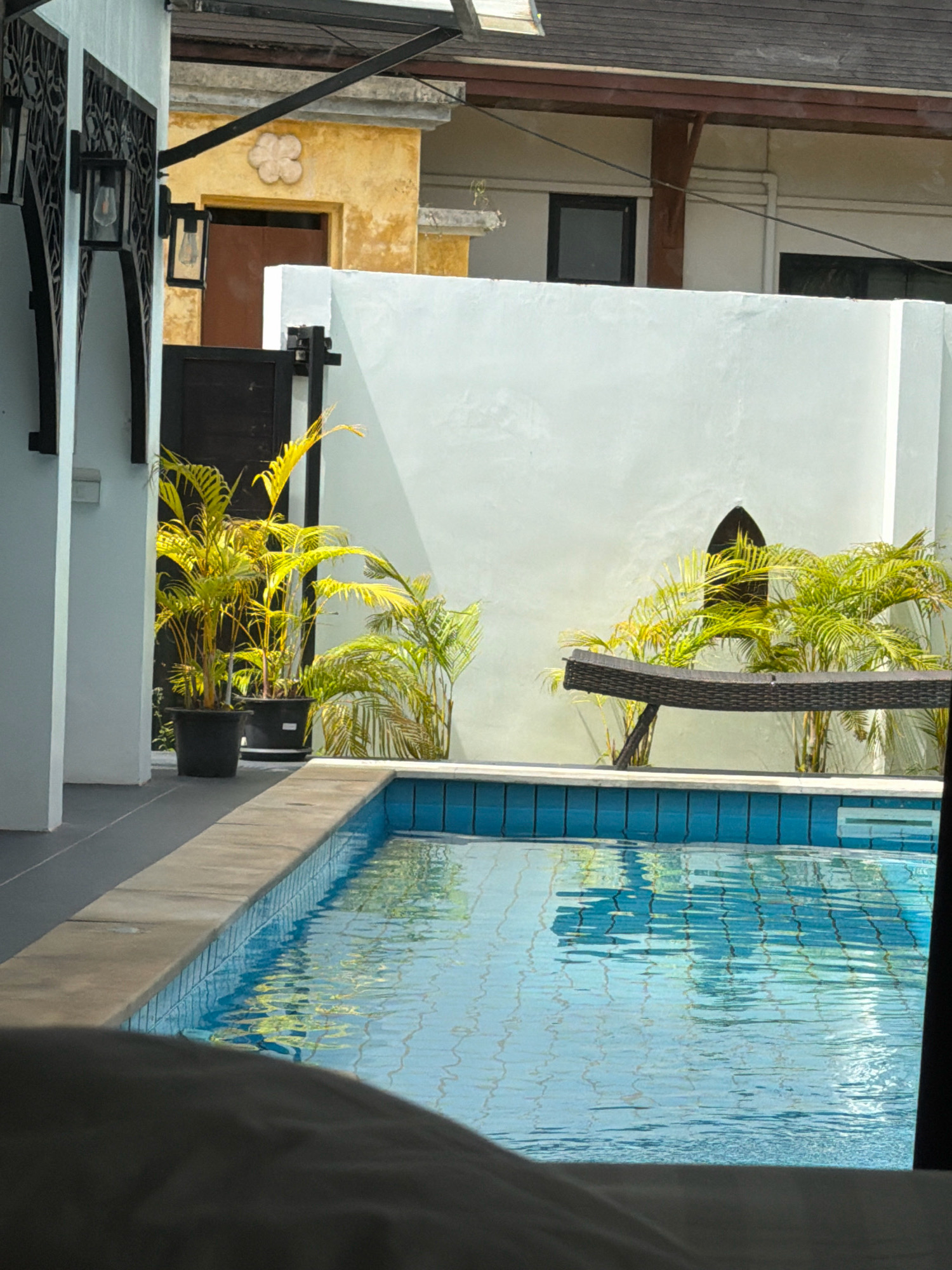 The image shows an outdoor swimming pool area in Si Sunthon, Thailand. In the foreground, a portion of a lounge chair is visible. The swimming pool itself is filled with blue water and tiled around the edges. To the left, there is a white wall with black decorative elements and wall-mounted lamps. Adjacent to the pool, on the left side, are two large potted plants with bright green foliage. In the background, there is a white wall with a dark, arched window-like feature. To the right of this wall, more greenery is visible. Further in the background, parts of buildings with yellow and white facades and a dark roof are seen. The scene appears to be a resort or private residence.