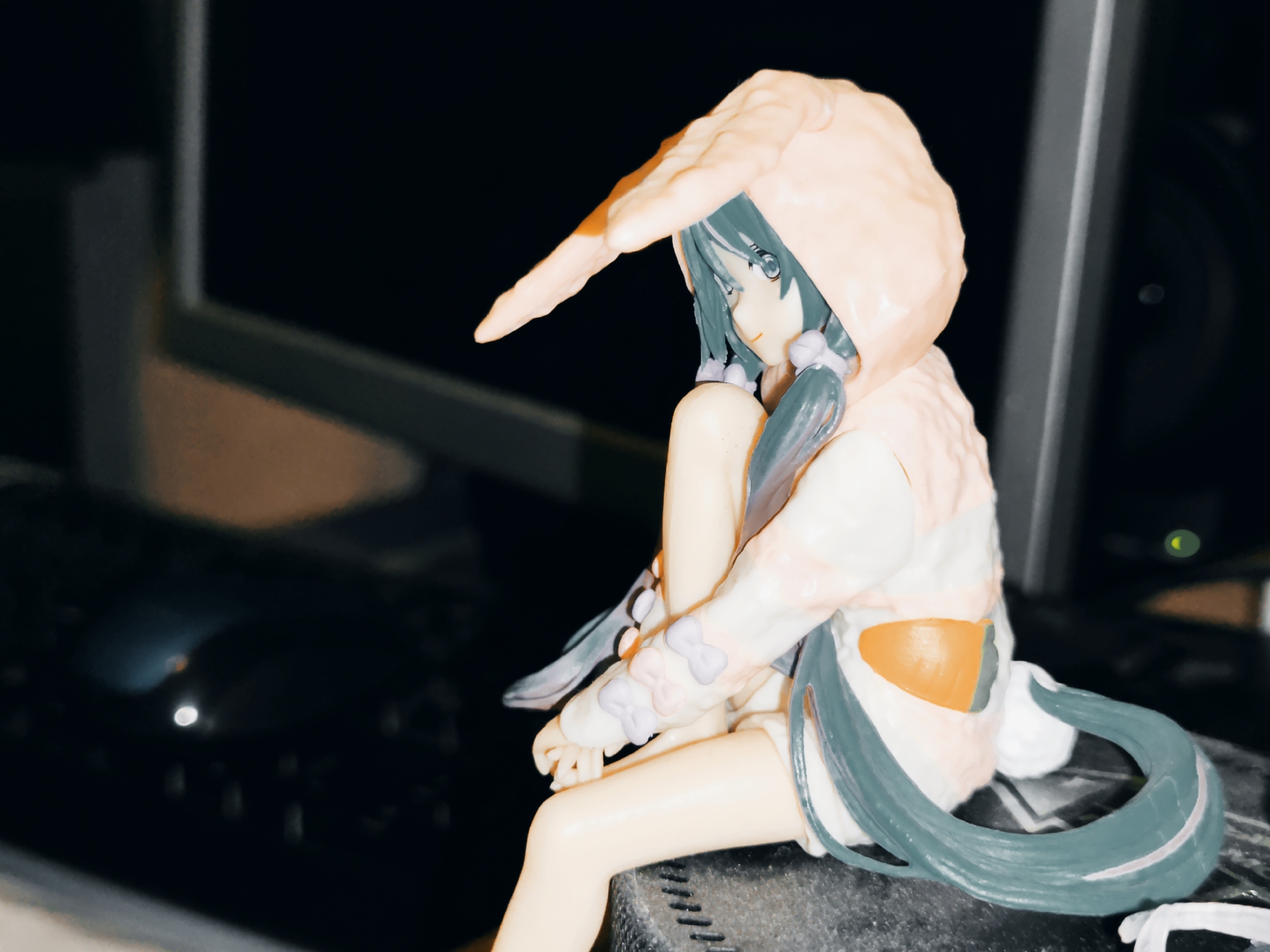 This media file features a close-up, side-profile shot of a figurine of a female anime character. The figurine is predominantly white and peach, with teal accents in her hair and clothing. She is wearing a hooded outfit with large, ear-like protrusions on the hood, resembling bunny ears. The character has teal hair styled in twin braids, and appears to be sitting with her legs crossed, her arms resting on her knees. Her expression is serene, with her eyes looking downwards and to the left. The figurine is positioned on a dark, textured surface, possibly a table or shelf. In the background, there is a dark, out-of-focus area that suggests an indoor setting, possibly with a screen or monitor visible. The lighting appears to be artificial and somewhat dim, creating a moody atmosphere. There is no visible text in the image. The location context is Makiivka, Ukraine.