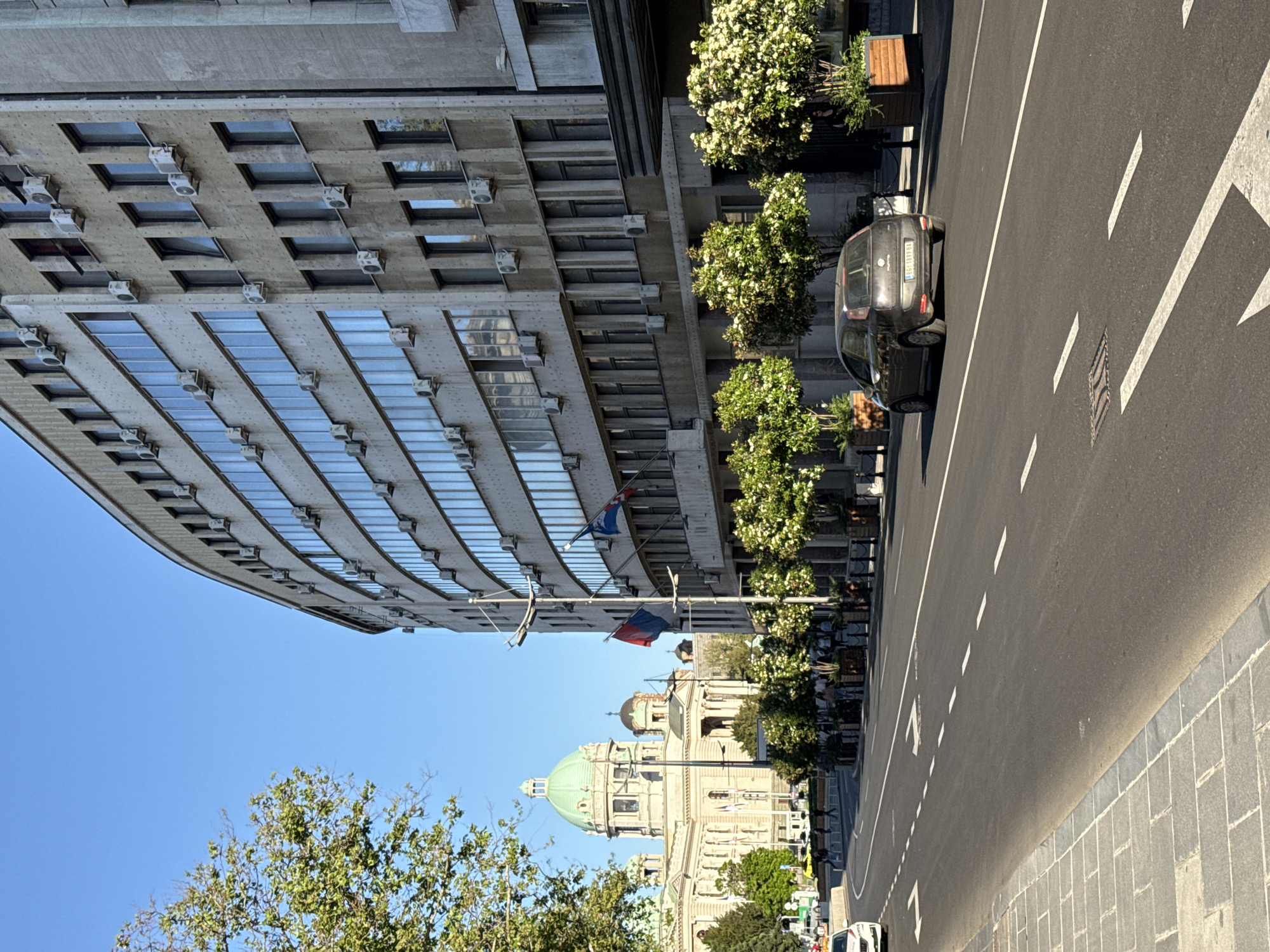The image shows a street scene in Belgrade, Serbia. A modern, curved building with numerous windows and air conditioning units is visible on the left side of the frame. A car is parked along the street, with decorative trees and planters positioned on the sidewalk near the building. Two Serbian flags hang from poles between the buildings. In the background, the dome of the Church of Saint Sava is visible. The street itself has multiple lanes with white lines marking the divisions, and is made of asphalt, while the sidewalk is constructed from gray stone tiles. The sky is clear and blue. Trees are also present in the foreground.