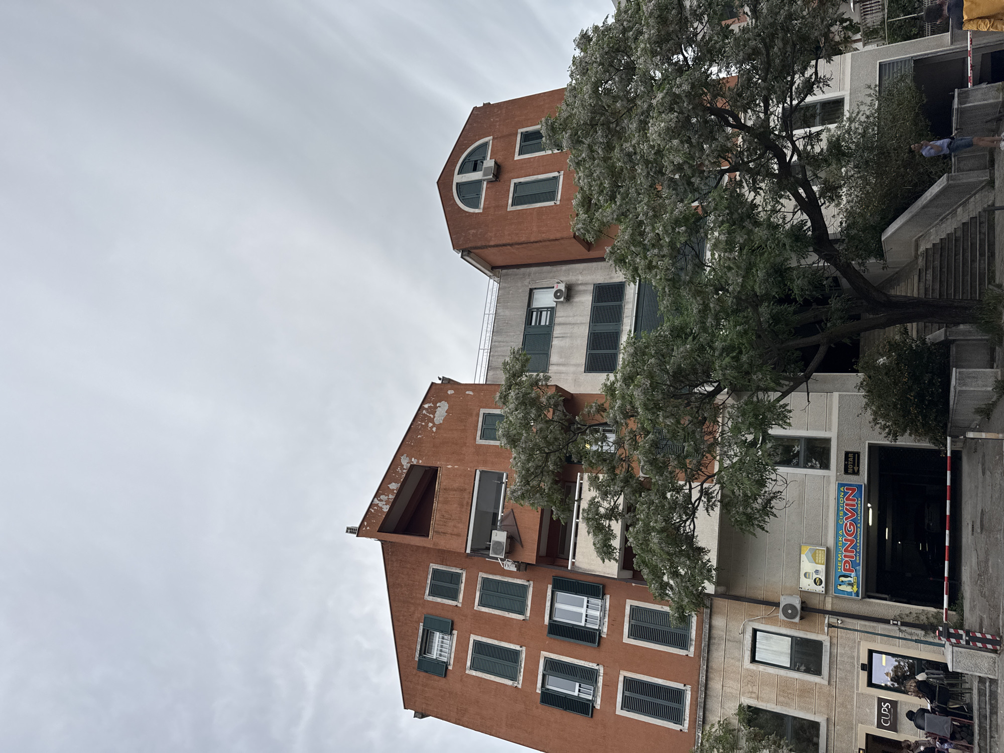 The image shows a building in Budva, Montenegro. The building is a combination of red-brown and white, with green shutters on the windows. The top of the building has a unique hexagonal shape with a semi-circular window. The building is adjacent to a commercial space with a sign that reads "HEMIJSKA CISTIONA PINGVIN". There is a tree in front of the building with white blooms. A person is walking up the stairs next to the building. There is a sky with a cloudy overcast appearance.