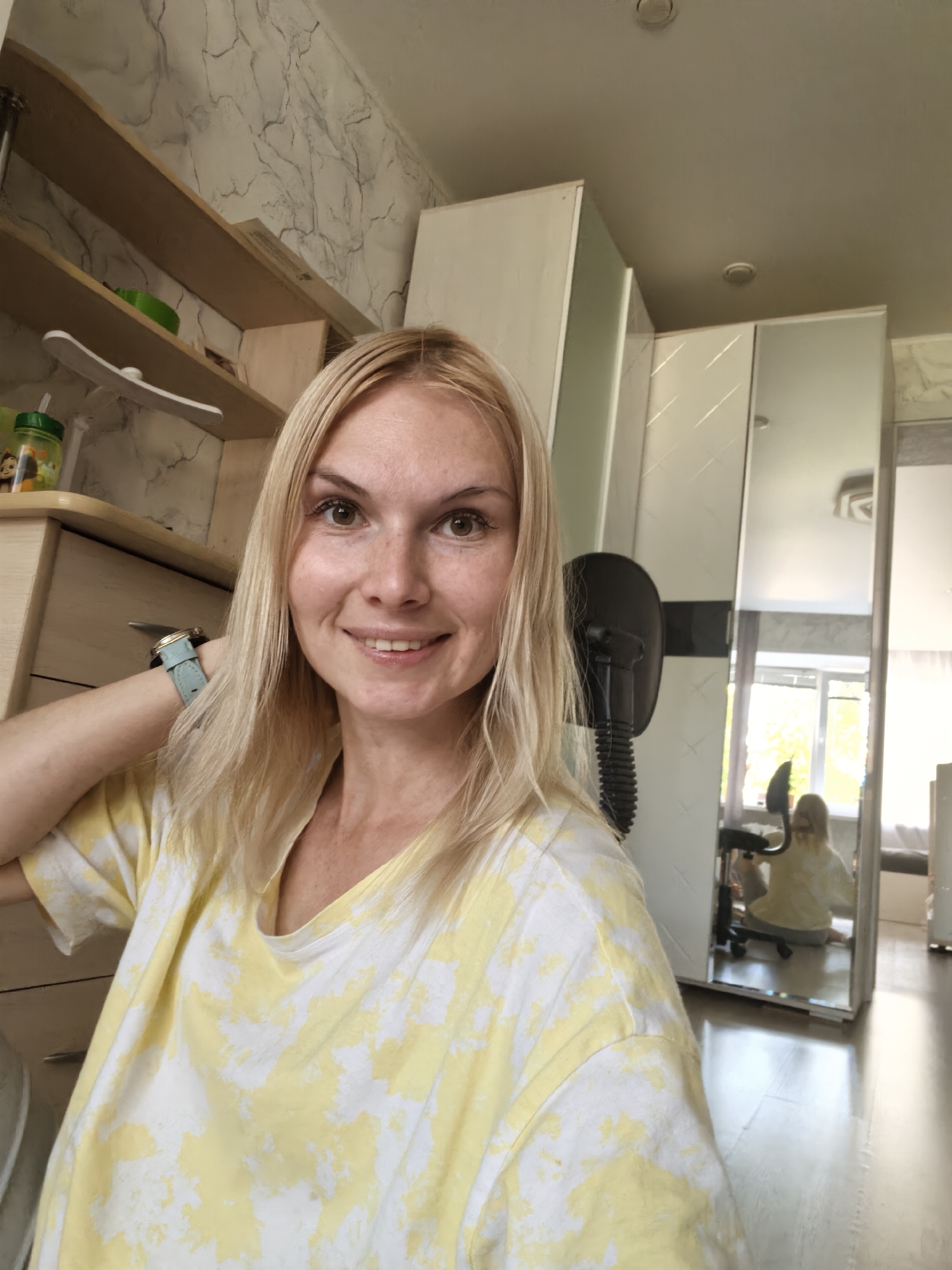 A blonde woman with fair skin and freckles is taking a selfie in a room. She is wearing a yellow and white tie-dye t-shirt. She is smiling and looking at the camera. Her left arm is raised, with her hand behind her head, revealing a light blue watch on her wrist. The background includes a wooden dresser, shelves with items on them, a large wardrobe with mirrored doors, and a desk chair. In the reflection of the mirrored wardrobe, another person, presumably the same woman, is visible sitting in the desk chair, facing away from the camera. The room appears to be a bedroom or living area, with a window visible in the reflection, suggesting daylight. The overall impression is of a casual, indoor setting. This image is not geotagged and cannot be associated with Sterlitamak, Russia.