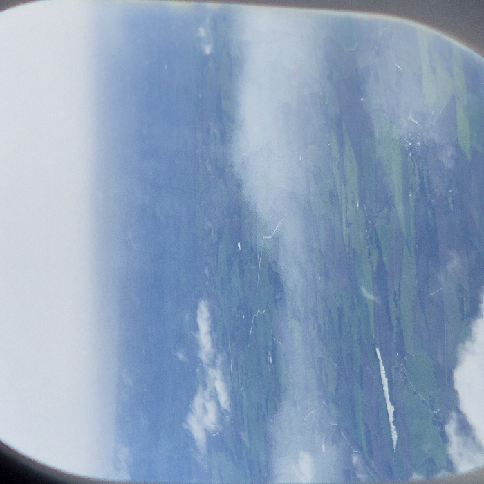 An aerial photograph, framed by an aircraft window, depicts a rural landscape observed from above, likely in the vicinity of Toulouse–Blagnac Airport near Blagnac, France. The scene features a mosaic of green agricultural fields of varying shades, traversed by multiple thin, light-colored roads or paths. A distinct, elongated body of water is visible in the lower-left section of the ground. Scattered white cumulus clouds partially obscure the terrain, with additional atmospheric haze present in the upper portion of the image. The horizon is indistinct. No individuals, discernible structures, or specific activities are identifiable.
