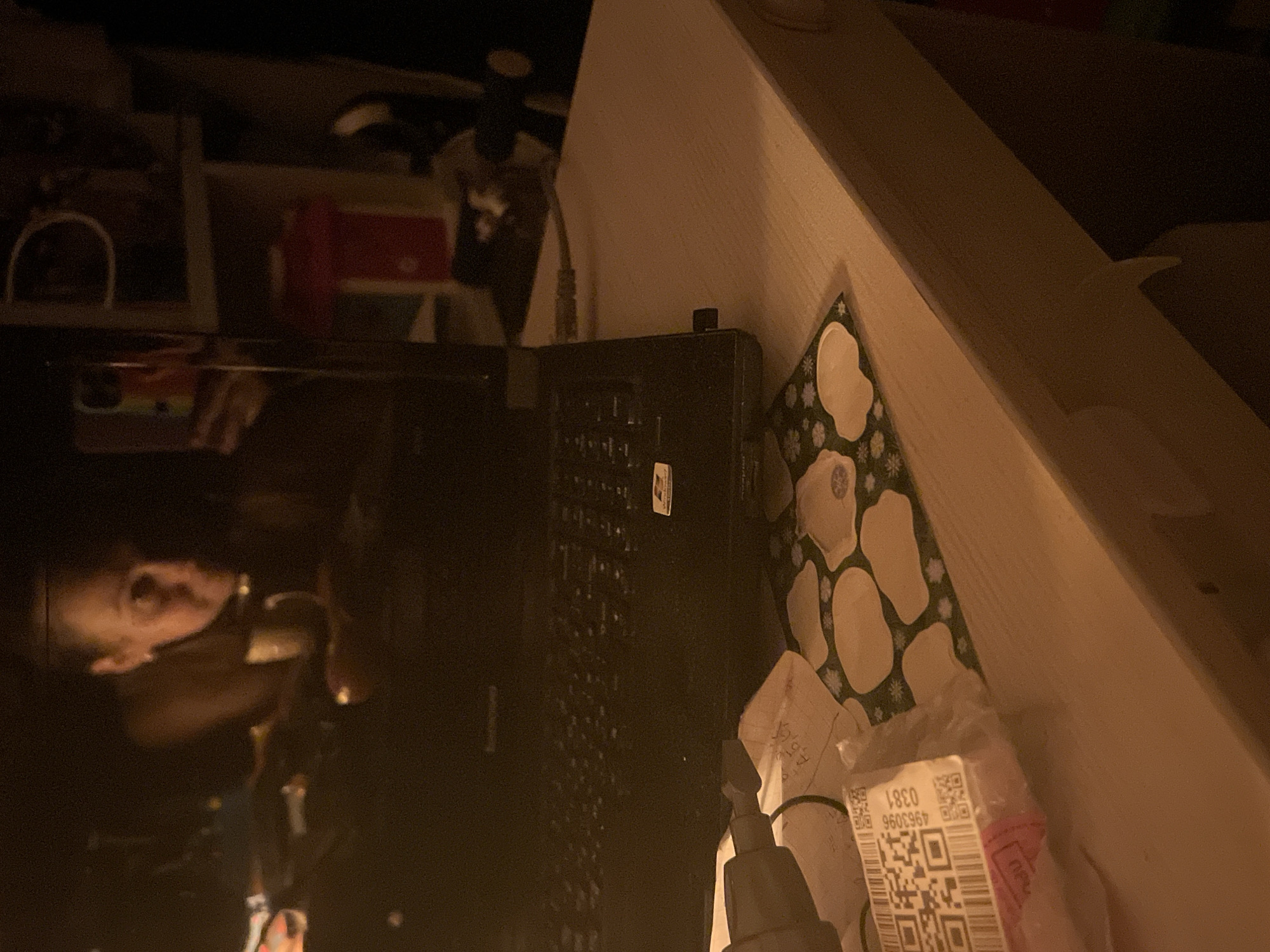 The image depicts a dimly lit indoor scene. A black laptop computer is prominently featured, positioned near the edge of a light-colored shelf or table. A reflection is visible on the laptop screen, possibly showing a person’s face. Adjacent to the laptop, there is a decorated surface, possibly a wrapping or a patterned item. A shipping label with a visible QR code is present, along with packaging material. The image does not provide specific evidence of the location being in Chaikovsky, Russia.