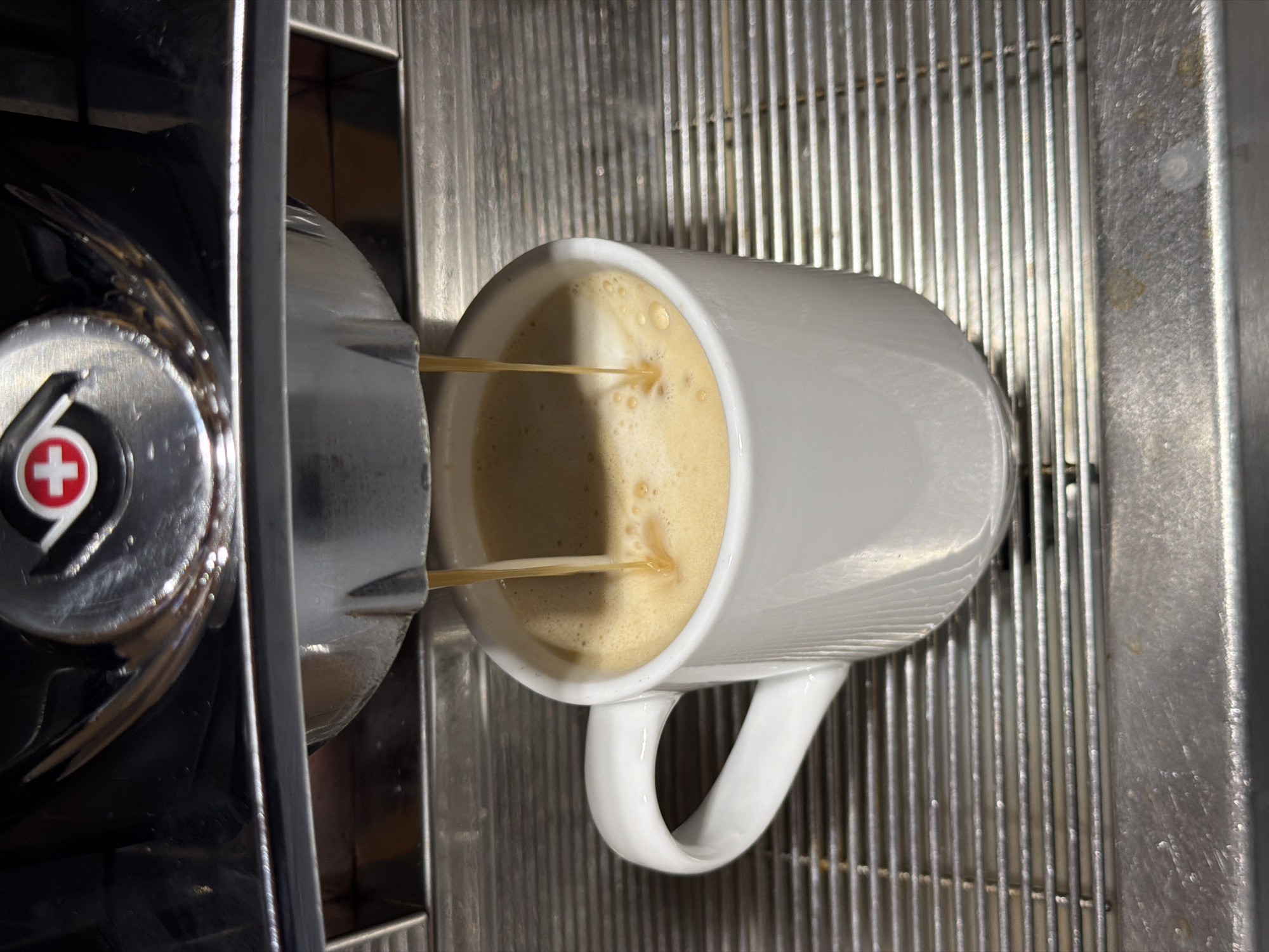 A white ceramic mug is positioned on a stainless steel grate beneath an espresso machine. Freshly brewed espresso is actively being dispensed into the mug, creating a layer of crema on top of the coffee. The espresso machine's chrome-plated dispensing head and a portion of its black casing are visible in the upper section of the image. A red and white cross emblem is visible on the espresso machine's surface. The surrounding environment appears to be indoors, likely a cafe or a home kitchen. The city of Garmisch-Partenkirchen, Germany, is not depicted or suggested in this image.