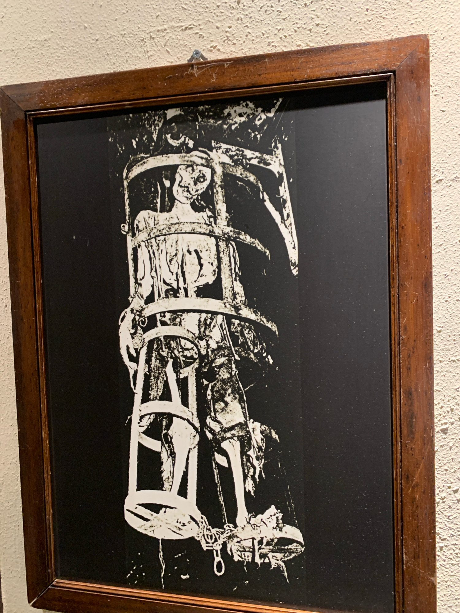 A framed black and white illustration depicts a skeletal figure in a cage. The figure appears gaunt and emaciated, with visible ribs and a skull-like head. It is confined within a cylindrical cage constructed of wooden bars and metal hoops. Chains are visible near the base of the cage, suggesting it may be suspended or capable of being moved. The background of the illustration is solid black, emphasizing the starkness of the figure and its confinement. The frame is made of dark wood with a visible grain and appears to be aged. The artwork is hung on a textured, cream-colored wall by a single screw.