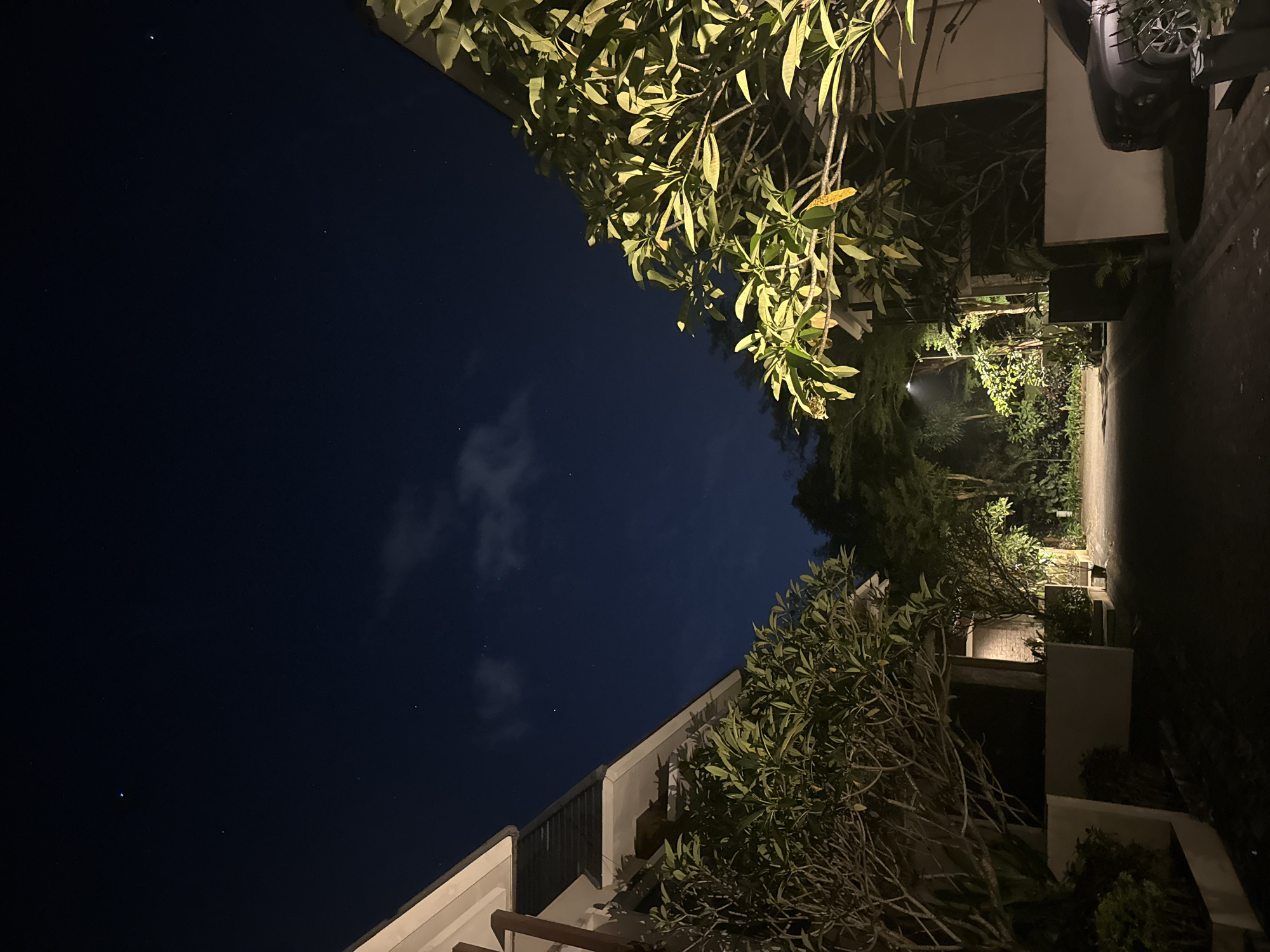 The image shows a nighttime scene in Kuta Selatan, Indonesia. A dark blue sky is visible above, with a few scattered clouds and stars. The foreground is framed by buildings and lush tropical vegetation, including trees with prominent leaves. The buildings are lit by warm artificial light, which also illuminates the foliage. A paved road or walkway leads into the distance, disappearing into the shadows further in. No people are visible. A car wheel is visible.