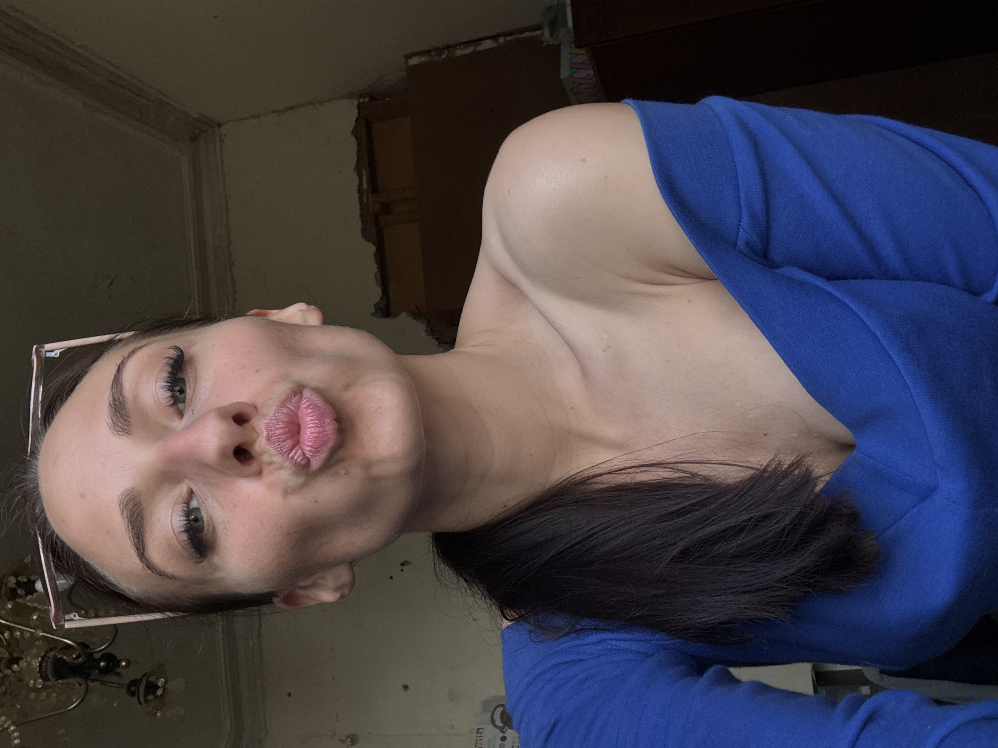 A young woman with dark hair pulled back in a ponytail is taking a selfie indoors. She is wearing a blue, off-the-shoulder top. Her lips are pursed in a kiss. She has glasses perched on her head. The background appears to be a room with peeling paint and an ornate chandelier. The context provided suggests this image was taken in Saint Petersburg, Russia, though there are no explicit visual cues in the image to confirm this location.