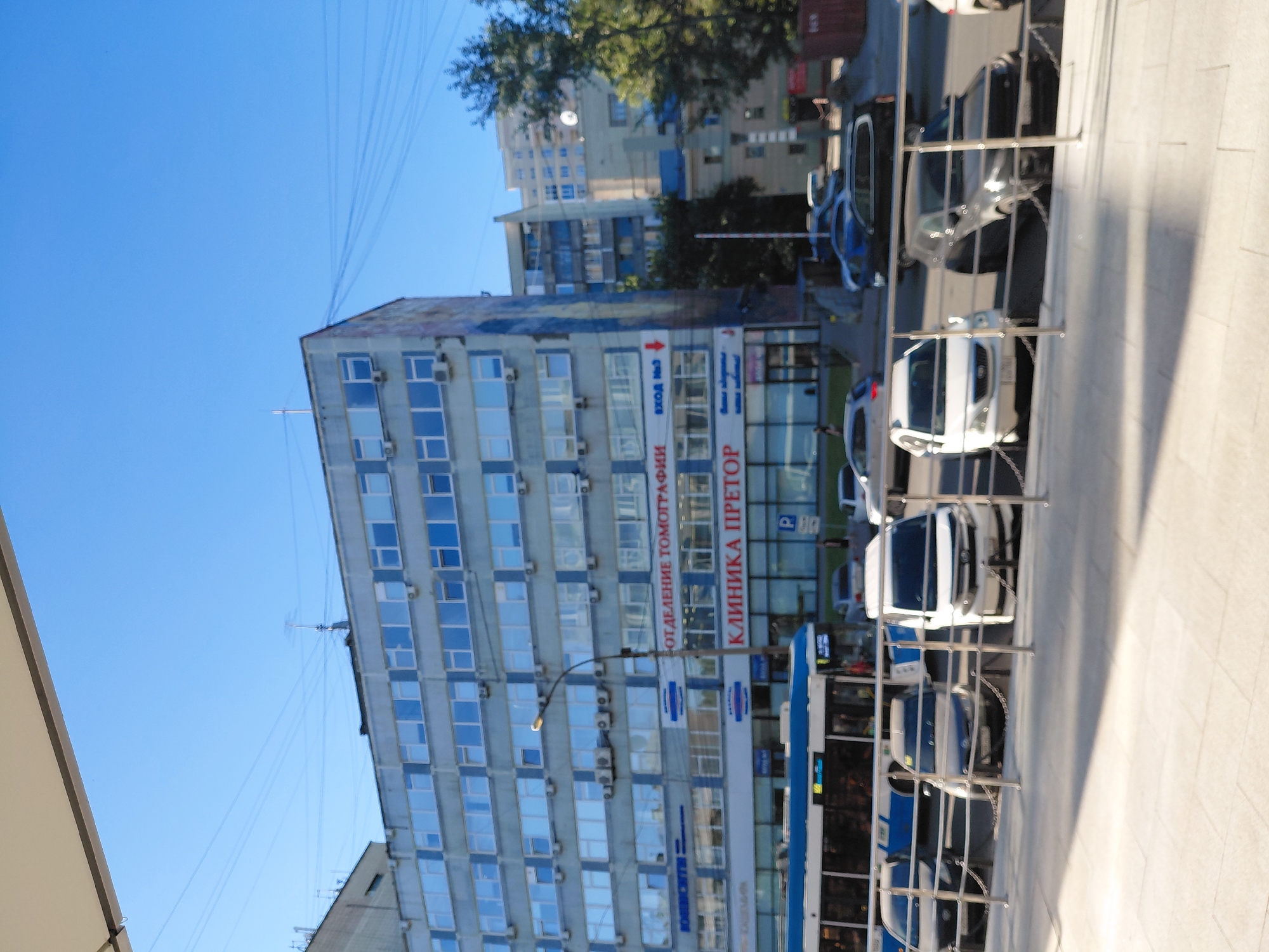 A wide shot captures a clear blue sky above a multi-story building in Novosibirsk, Russia. The building, with its pale blue facade and numerous windows, dominates the frame. A bus with a blue and white exterior is parked in front of the building, alongside several cars. The cars are parked behind a metal railing. A street lamp stands between the building and the street. The ground in the foreground is paved with light-colored tiles. The overall scene is one of an urban environment bathed in sunlight.