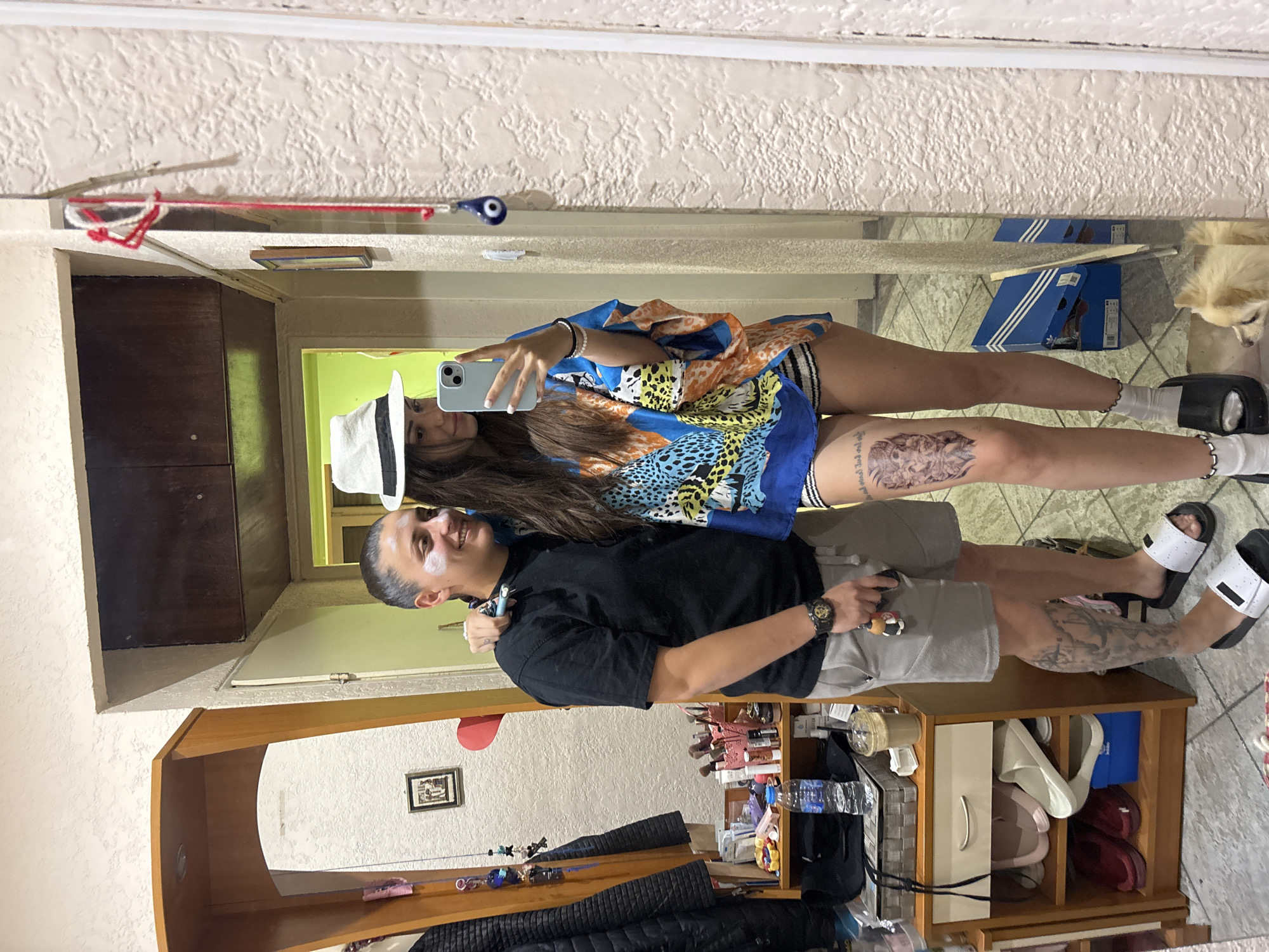 Two individuals are taking a mirror selfie indoors. On the left, a person with short, dark hair and facial cream applied to their cheeks and forehead wears a black t-shirt, grey shorts, and white slides with black accents. They are wearing a watch on their left wrist and hold a small keychain. On the right, a person with long, dark hair and a white fedora with a black band wears a colorful, loose-fitting top with a leopard print pattern, black and white striped shorts, and black platform slides with white accents. They are wearing white ankle socks with decorative rings around the top. A large tattoo of a face is visible on the right person's left thigh. Behind them, a shelf unit with various items, including shoes, toiletries, and a bottle of water, is visible. A small, light-colored dog is lying on the floor in the lower right corner. The overall setting appears to be a residential interior with textured walls and a tiled floor.