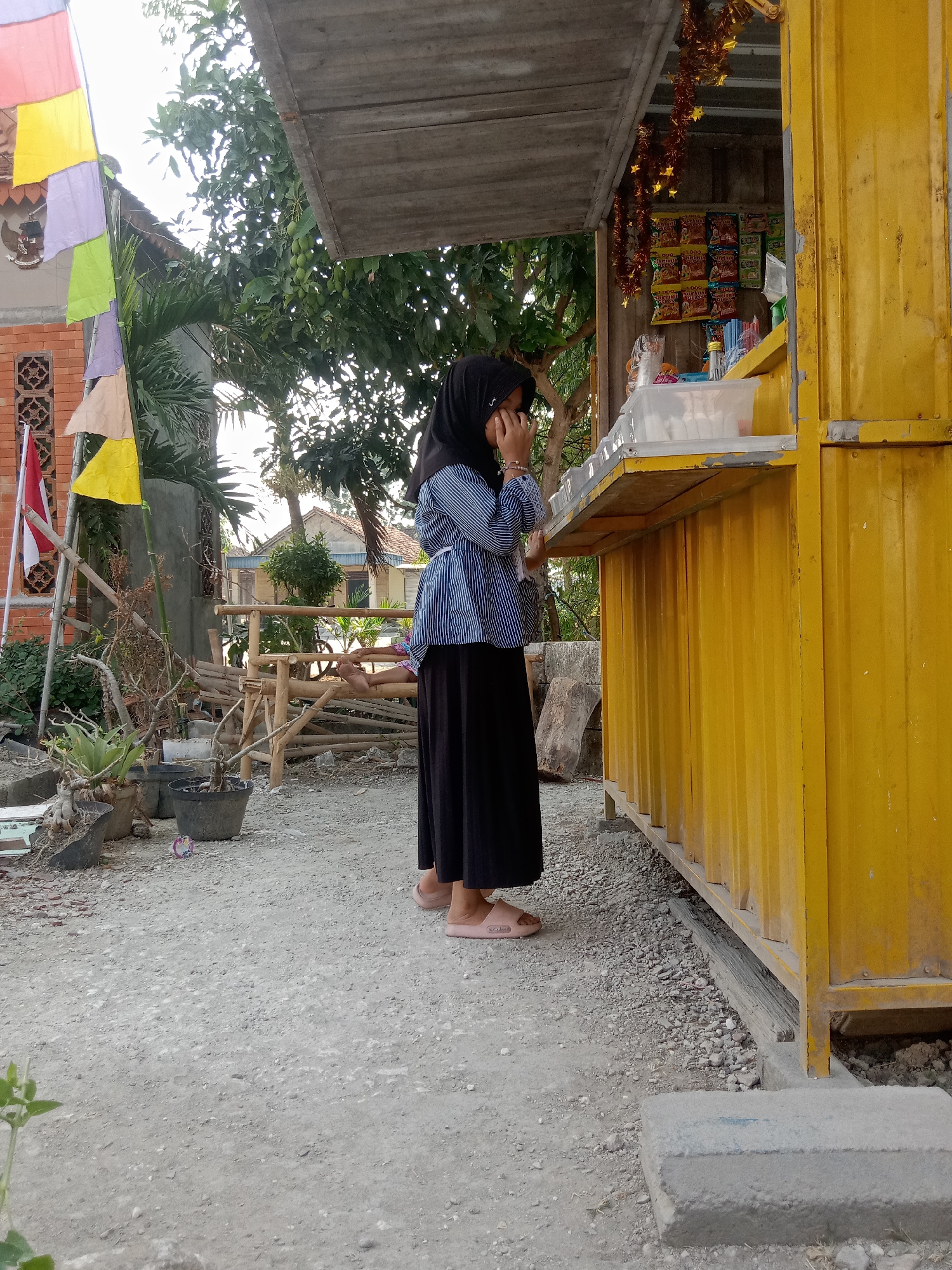 A young girl wearing a black hijab and a blue and white striped shirt stands facing a yellow kiosk in Stiaji, Indonesia. She is covering her face with her hands, and her pink sandals are visible. The kiosk appears to be a small shop selling snacks, with shelves stocked with packaged goods. The ground is covered in gravel and pebbles, and there are some potted plants and wooden structures nearby. In the background, a building with a red brick wall and flags is visible, along with trees and houses. The scene suggests a casual outdoor setting, likely in a residential or commercial area.