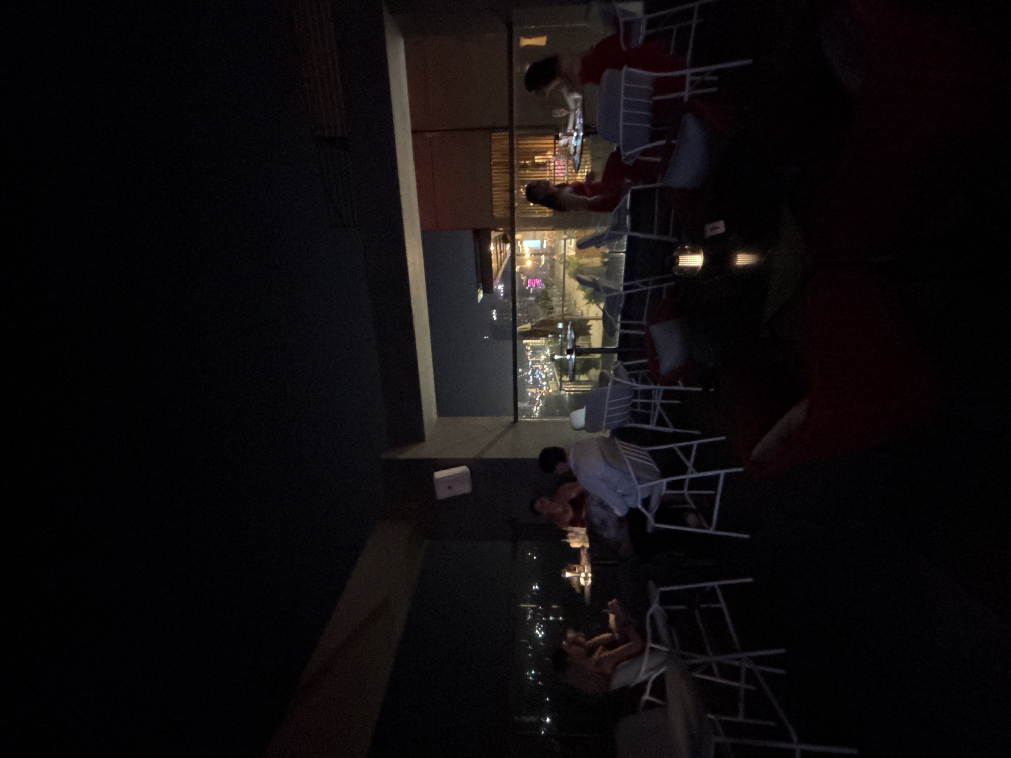 The image depicts a rooftop bar at night in Da Nang, Vietnam. The scene shows several groups of people seated at tables, with a city skyline illuminated in the background. In the foreground, two women in red dresses are seated at a table with glasses, possibly enjoying drinks. To the left, other patrons are visible, some engaged in conversation. The bar area features modern white chairs and tables, and ambient lighting from small lamps on the tables and distant city lights. The overall atmosphere suggests a social gathering or dining experience in a contemporary urban setting.