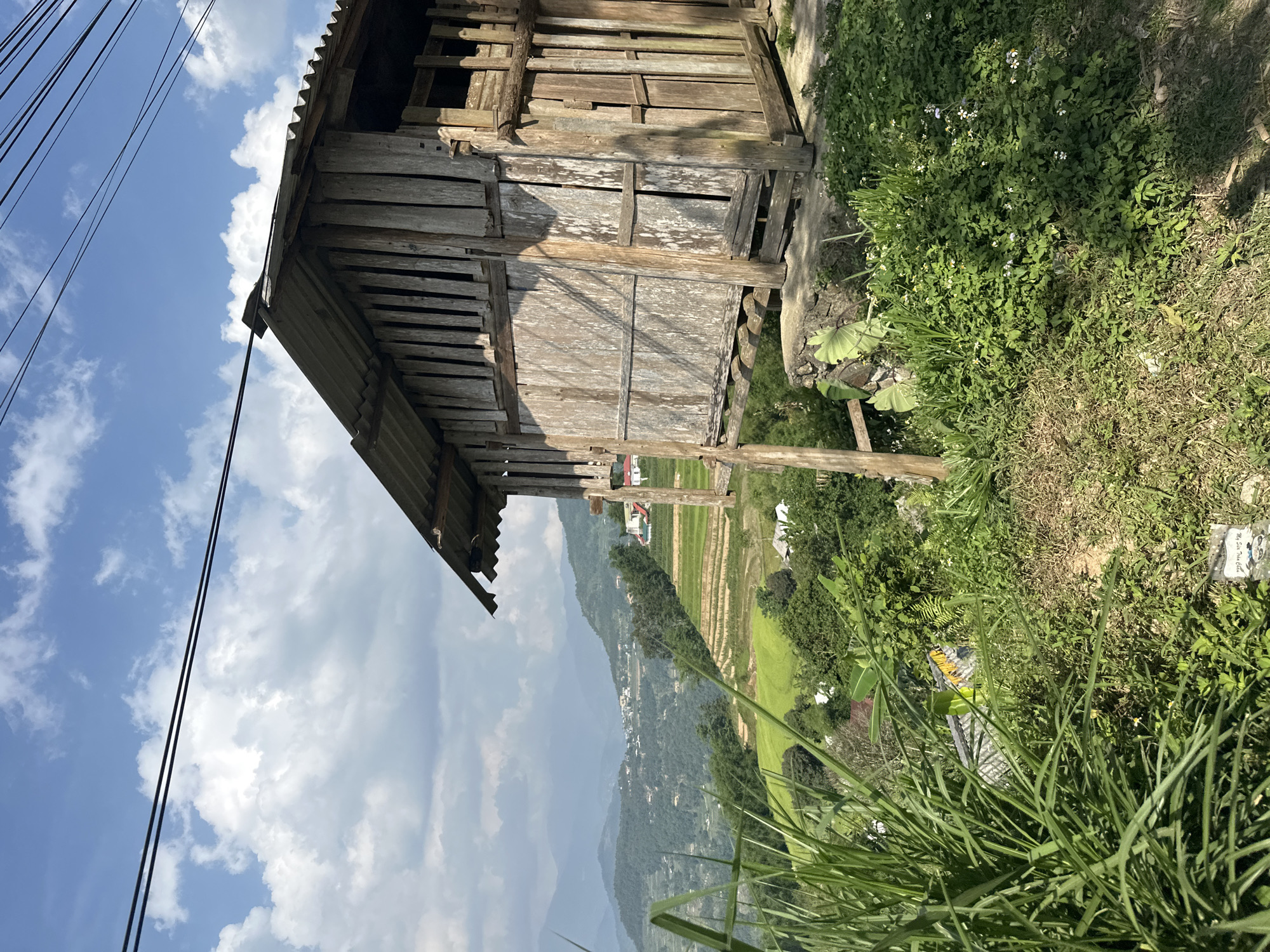 An elevated, tilted wide shot of a wooden structure in Pờ Ly Ngài Commune, Vietnam. The structure, possibly a shed or small house, is made of weathered wooden planks and has a corrugated metal roof. Part of the structure's wall is open, revealing the interior framework. Adjacent to the structure, the landscape slopes downward covered in dense green vegetation, including grass and small flowering plants. In the background, terraced rice fields are visible, cascading down the hillside. Further in the distance, mountains fade into a cloudy sky. Black power lines are visible against the sky.