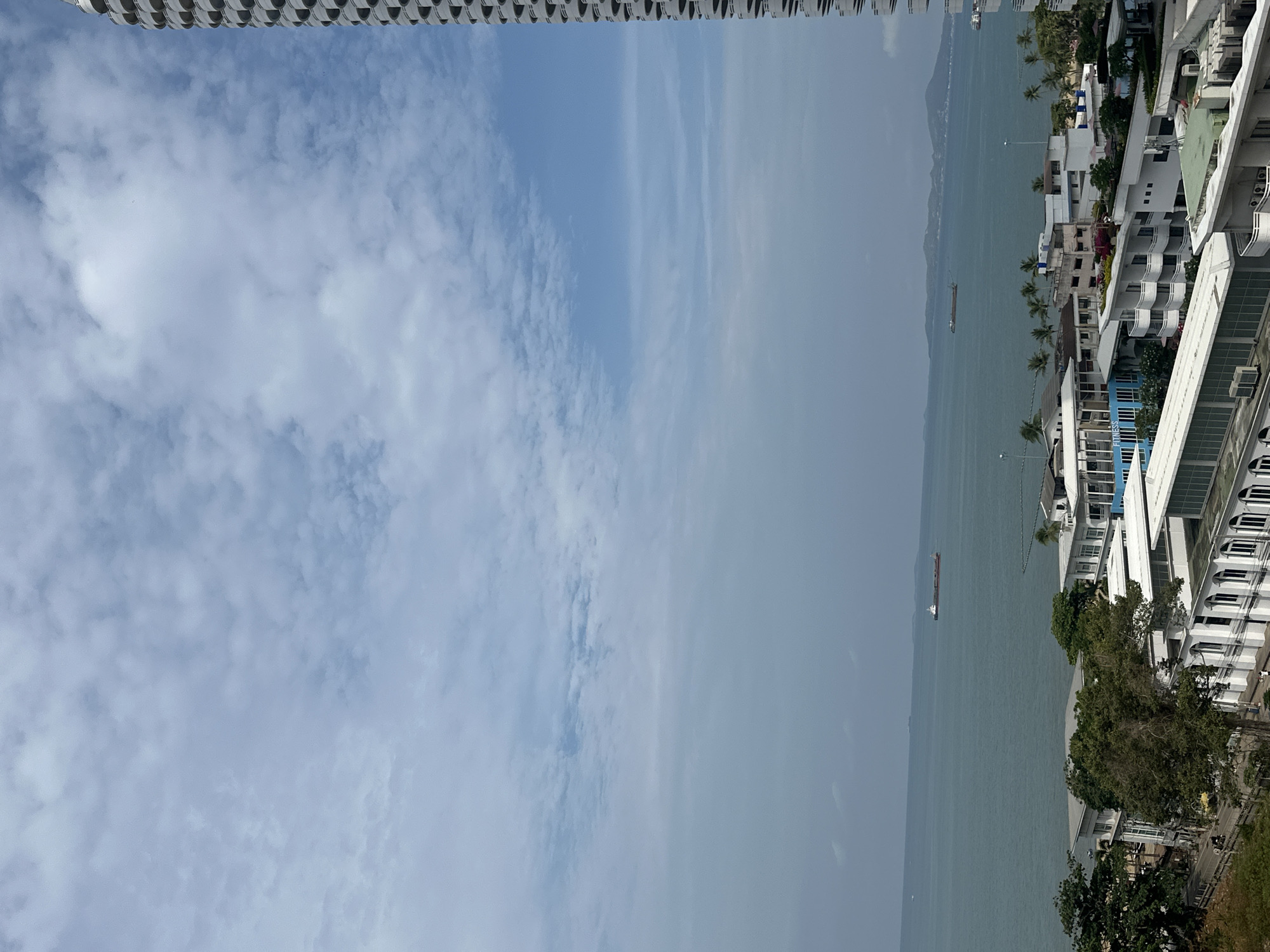 The image is an outdoor shot of Pattaya City, Thailand, taken from an elevated position, possibly a high-rise building.
The sky is mostly cloudy, with shades of blue visible amidst the clouds. A portion of a tall building, possibly the photographer's location, is visible at the top of the frame.
A body of water stretches horizontally across the midsection of the image. Several boats and ships are visible on the water.
The right side of the image shows a dense area of buildings, with a mix of residential and commercial structures. Trees and greenery fill the foreground.