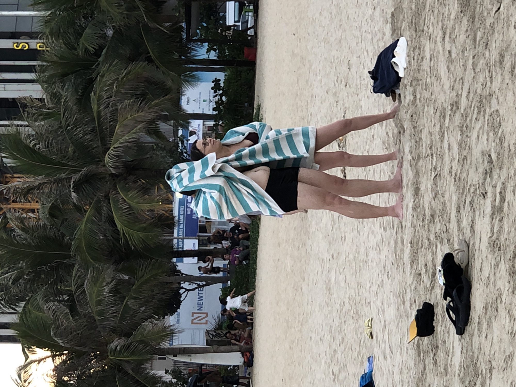 Two individuals, one appearing to be a woman wearing sunglasses and a bikini top, and the other a person in black swim shorts, are standing on a sandy beach. They are partially covered by a large, striped towel. In the background, palm trees are visible, along with buildings and what appears to be signage in Vietnamese. Scattered on the sand are various objects, including clothing, flip-flops, and a yellow object that might be a child's toy. In the mid-ground, a group of people are sitting on the sand and grass, some appearing to be observing the scene. The overall impression is of a casual beach setting, possibly during the late afternoon or early evening, given the lighting. The presence of Vietnamese text on the signage strongly suggests the location is in Vietnam, specifically Đà Nẵng. The people on the beach appear relaxed and engaged in typical beach activities.