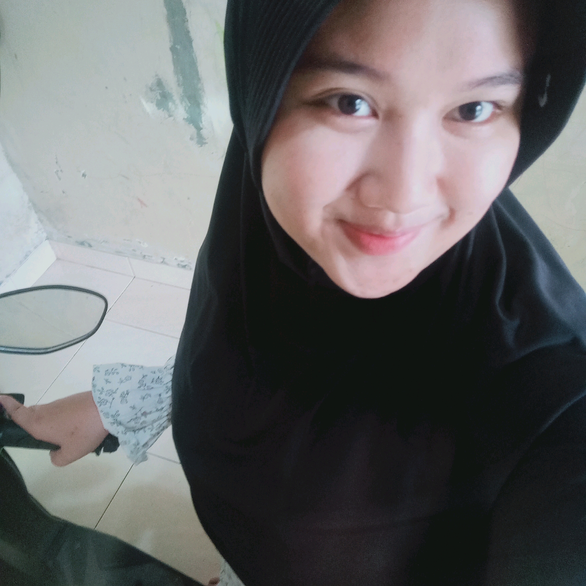 In Purwakarta, Indonesia, a young woman wearing a black hijab is captured in a selfie. She is seated, likely on a motorcycle, as evidenced by the handlebars and a side mirror visible in the lower left of the frame. The background consists of a tiled floor and a light-colored wall with some discoloration and texture. The woman's face is framed by the hijab, and she has a gentle smile. Her left arm is extended, and her hand is resting on the handlebars. She is wearing a light-colored blouse with a subtle floral pattern. The image has a casual, personal feel, typical of a self-taken photograph.