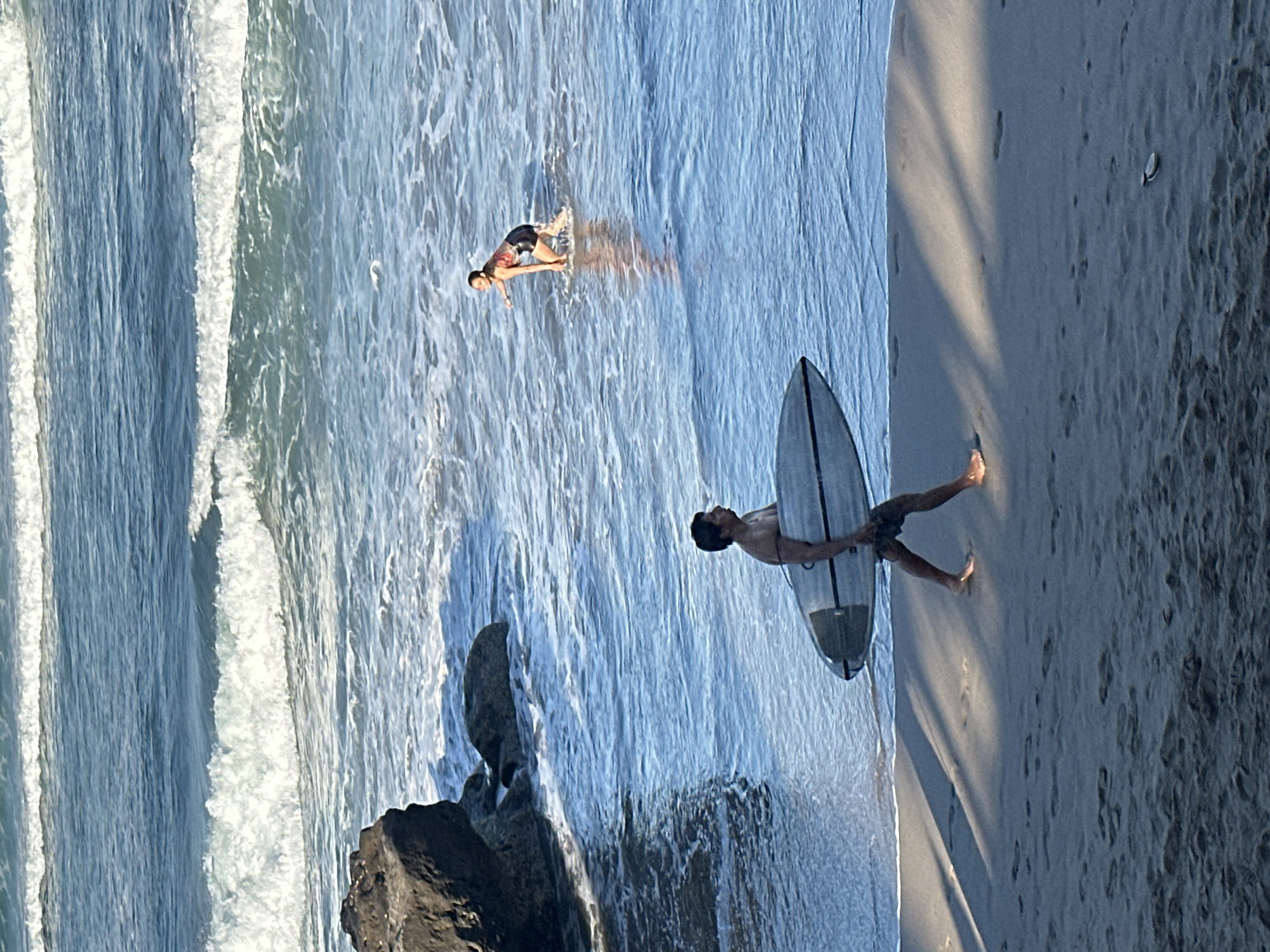 The image depicts a beach scene in North Kuta, Indonesia. In the foreground, a shirtless man with dark hair walks along the sandy shore carrying a surfboard. He is wearing dark shorts. The surfboard is light-colored with dark accents. His legs are casting shadows on the sand. Further out in the shallow water, a woman with dark hair is crouched, facing away from the camera and to the left, with her right arm extended as if pointing or reaching. She is wearing a dark-colored top with a patterned design and dark shorts. The ocean is blue with breaking waves in the background, and some dark rocks are visible on the left side of the image near the water's edge. The lighting suggests it is daytime, possibly late afternoon due to the long shadows.