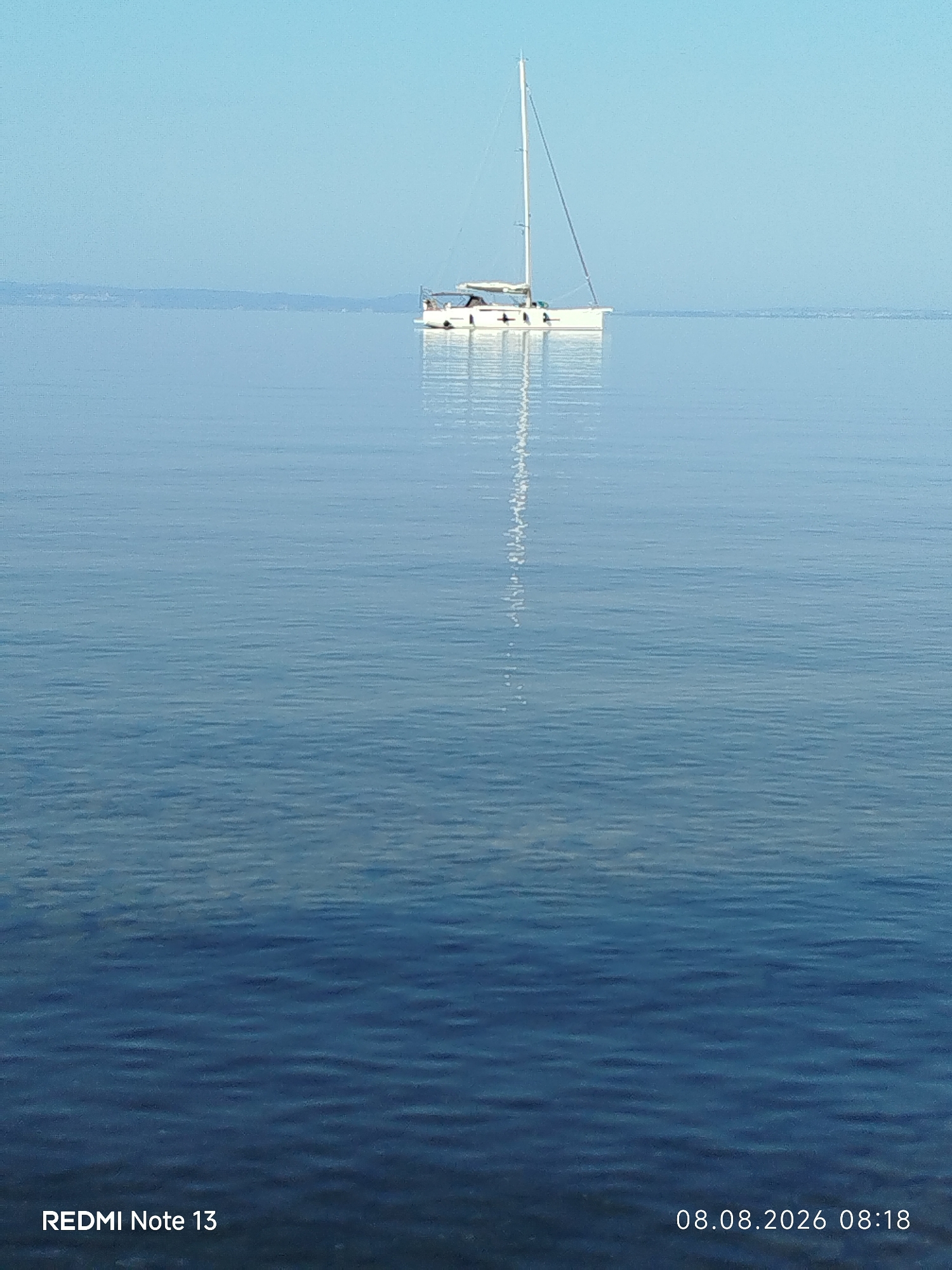 A lone white sailboat with a tall mast is at anchor on calm, blue water. The boat has a small cabin with windows and appears to have at least two people visible on deck. A faint reflection of the boat and mast is visible on the water's surface. In the distance, a hazy landmass, likely the coastline of Sithonia, Greece, is visible against a clear, pale blue sky. The bottom left corner of the image displays the text "REDMI Note 13", and the bottom right corner shows the date and time "08.08.2026 08:18". The water in the foreground is a darker shade of blue than the distant water.