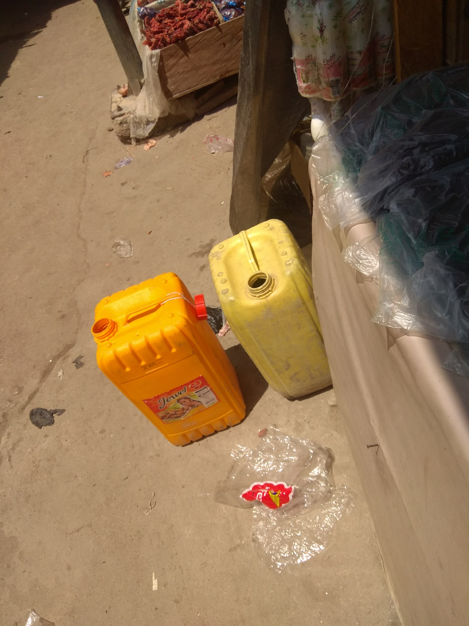 In Jalingo, Nigeria, the scene is set on a sun-drenched outdoor surface, likely a market or roadside stall, with a rough, concrete ground. The primary subjects are two large plastic jerrycans, one bright orange and the other a dull yellow, both appearing to be empty. The orange jerrycan has a label that reads "Jewel" in prominent red lettering, with a depiction of food items on it. A red cap is visible on its spout. The yellow jerrycan is positioned behind and to the right of the orange one, and its spout is open. Scattered around the jerrycans are pieces of clear plastic trash, one with a red and yellow logo, suggesting an area with commercial activity.
In the background, a wooden crate overflows with a mound of dried red chili peppers, indicating produce being sold. Further back, draped fabrics or plastic sheeting, possibly covering other goods, are visible. The lighting suggests it is daytime, with strong sunlight casting shadows and highlighting the textures of the ground and the plastic containers. The overall impression is one of a typical, bustling marketplace environment, possibly in a warm climate. No people are directly visible in the frame, but their presence is implied by the commercial setting. The scene is devoid of any overt emotional cues, presenting a straightforward depiction of everyday objects in a specific location.