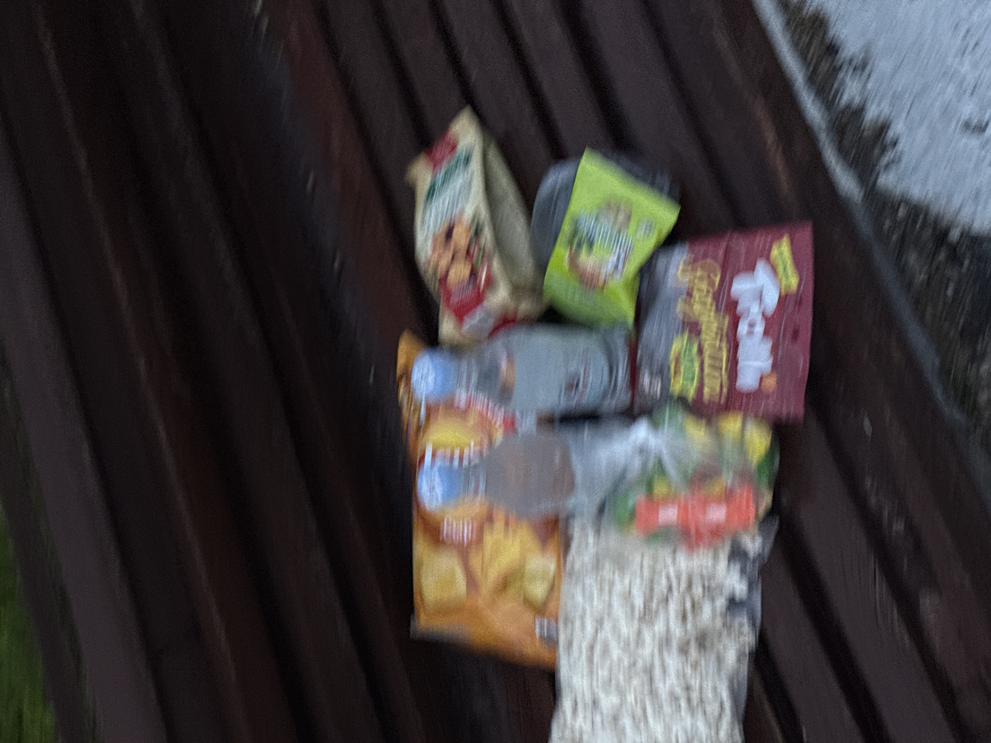 The image shows a collection of snack food packaging and what appears to be cooked noodles, possibly leftovers, placed on a dark brown slatted surface, likely a bench. The specific brands and varieties of the snacks are difficult to discern with clarity due to the image's blurriness, but there are chip bags, candy wrappers, and potentially other processed food items. The setting implies a public space, and the prompt suggests the location is in Yerevan, Armenia, without more details is difficult to provide further forensic information. There are no people visible in the image.