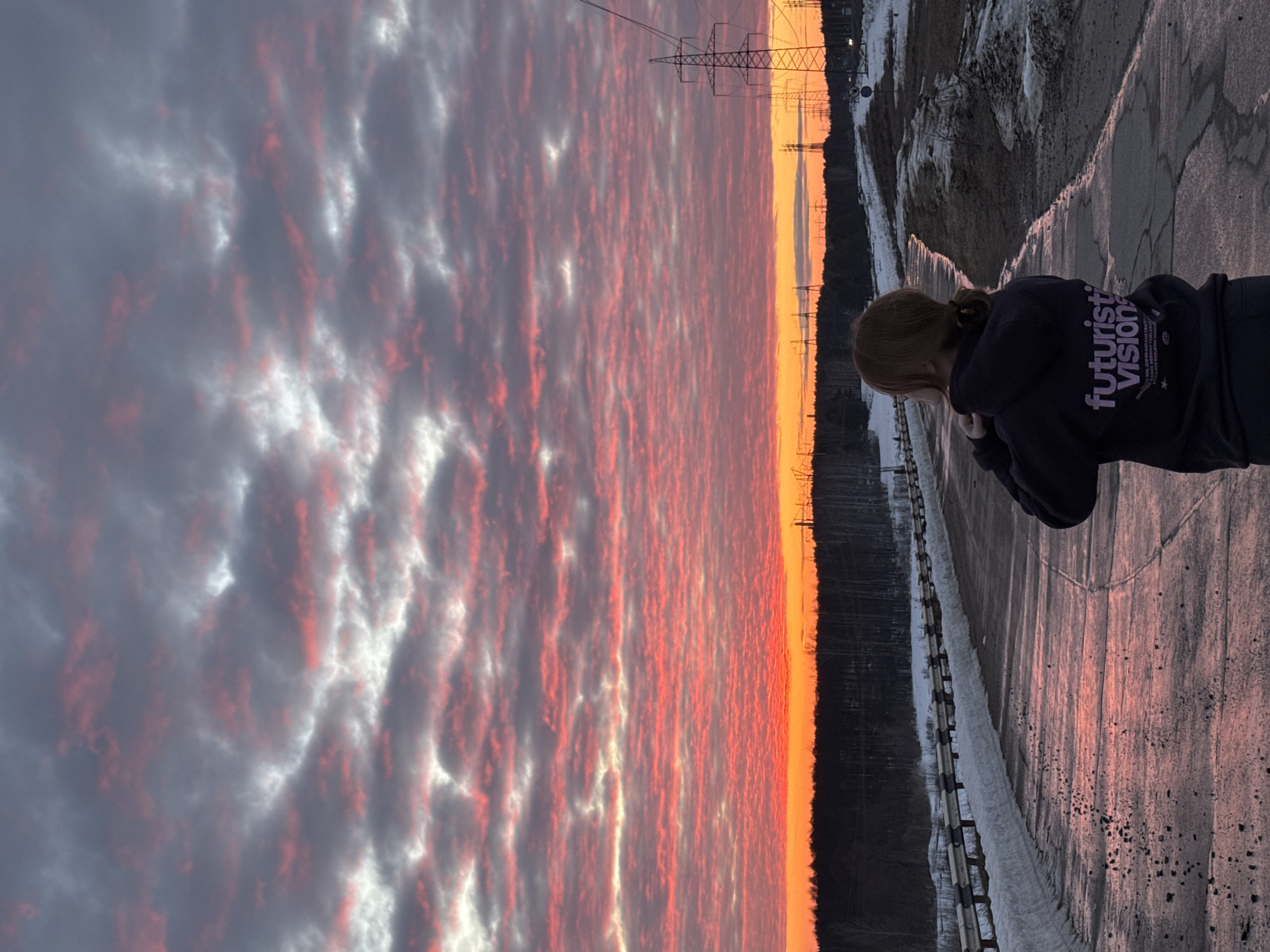 An individual stands with their back to the viewer on a wet, reflective asphalt road on the outskirts of Chaikovsky, Russia, facing a sunset. The person has reddish-brown hair pulled into a ponytail and is wearing a dark hooded sweatshirt featuring "futuristic visions" printed in purple text on the reverse. The road surface is wet, reflecting the sky's colors, and is dotted with scattered dark debris. To the immediate left, a metal guardrail runs parallel to the road, bordered by a continuous accumulation of snow. To the right, the road's edge transitions into a dirt and snow-covered shoulder. In the background, a dark line of bare forest trees is silhouetted against the horizon. Multiple electricity transmission pylons are visible along the right side of the horizon, extending into the distance. The sky above is dominated by a sunset, displaying a bright orange band at the horizon, which transitions upwards into a widespread formation of altocumulus clouds illuminated in intense red and pink hues, interspersed with patches of grey and white.