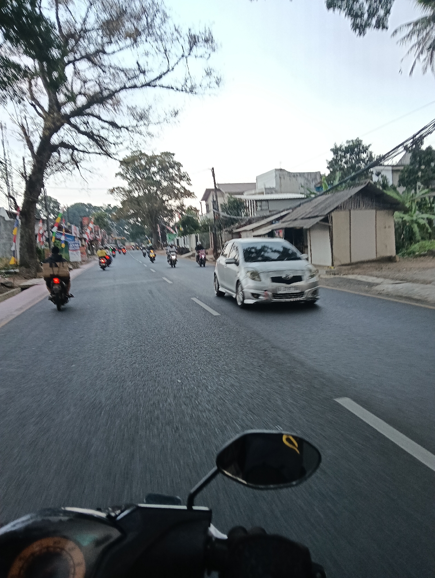 This is a photograph taken from a motorcycle, looking forward on a road in Tasikmalaya, Indonesia. The sky is overcast and appears to be either early morning or late afternoon. The road is paved and has white lane markings. Several motorcycles and a silver car are visible on the road. On the left side of the road, there are trees, buildings, and colorful flags. The foreground of the image shows the handlebars and dashboard of the motorcycle from which the photo is taken. The overall impression is a busy street scene in an urban environment.