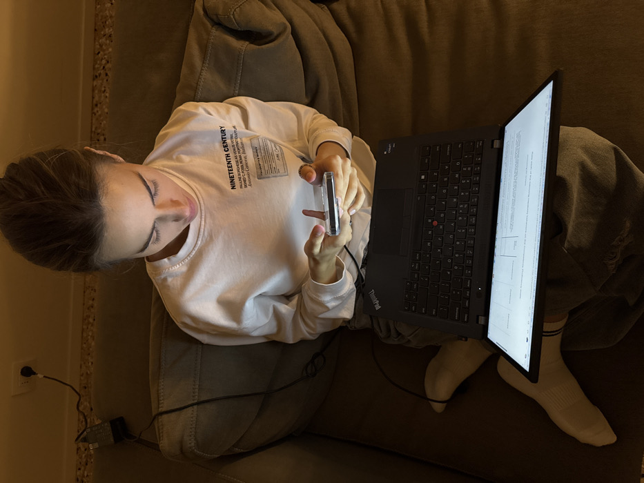 A young adult female is depicted from an elevated perspective, seated on a dark olive green or brown fabric couch in an indoor setting within the city of Venice, Italy. She wears a light-colored, long-sleeved sweatshirt, featuring text on the chest that reads "NINETEENTH CENTURY" and a label-like graphic beneath it, which includes the phrase "PAULINE BORGHESE AS VENUS VICTRIX." Her hair is pulled back, and she is wearing light-colored pants, possibly distressed jeans, and white socks.
She is actively engaged with two electronic devices. In her hands, she holds a smartphone with a clear case, its screen illuminated, and her fingers are positioned as if typing or navigating. A black Lenovo ThinkPad laptop rests open on her lap, its screen also illuminated, displaying what appears to be a document or form with visible text.
The immediate environment is dimly lit by warm artificial light, predominantly from the screens of the devices. A black power adapter is plugged into a wall socket (Type F or L, common in Italy) on a light-colored wall directly behind the couch, with a cable extending downwards, likely connected to one of the electronic devices. The overall scene suggests a casual, focused activity in a residential space.