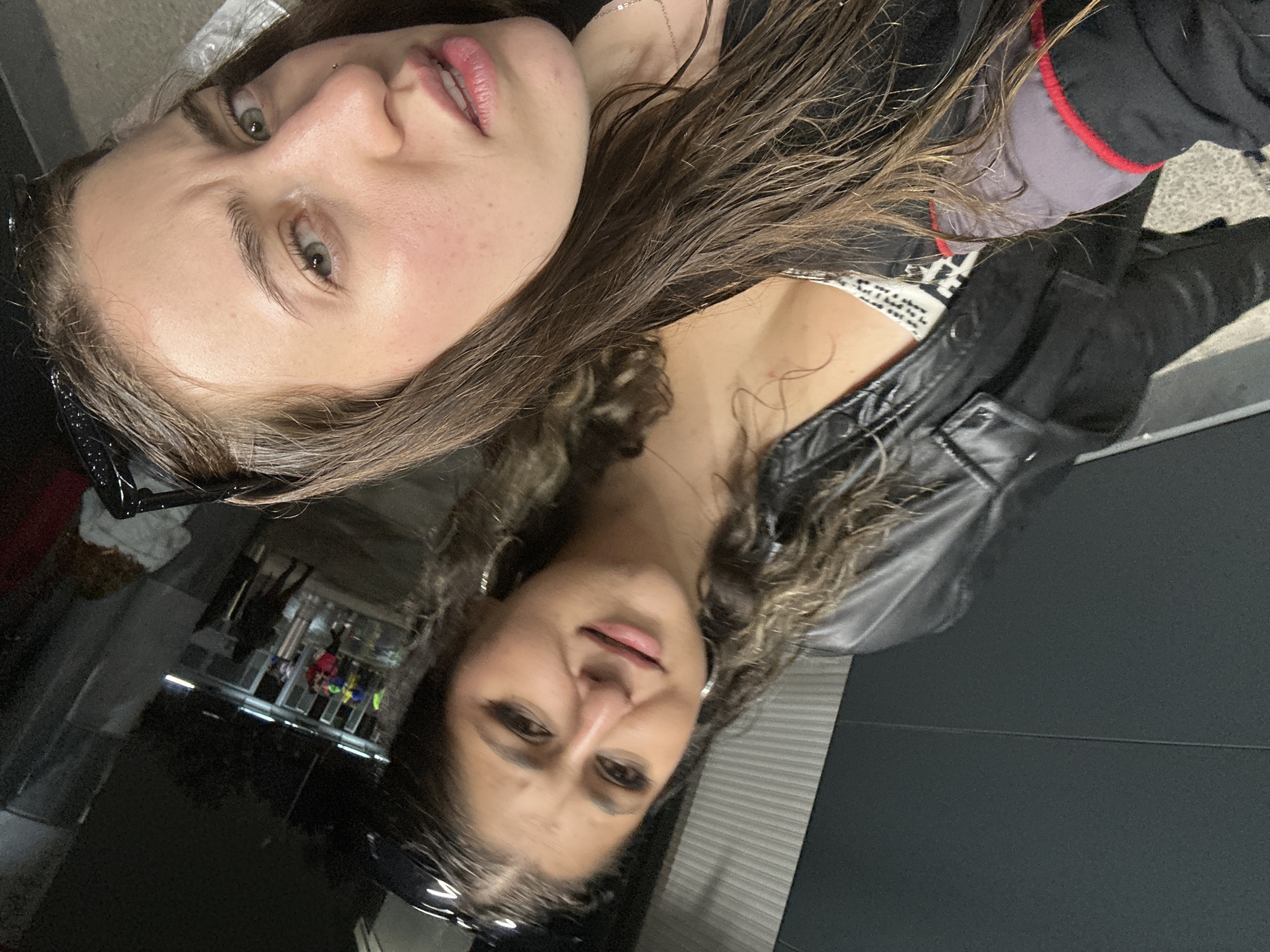 Two women pose closely together for a photograph. The woman on the left has long brown hair, fair skin, and a nose piercing. The woman on the right has long brown curly hair and wears a black leather jacket. The setting appears to be in Mexico City, possibly in an indoor urban environment, such as a transportation hub, as suggested by the storefronts reflected in the glass behind them.