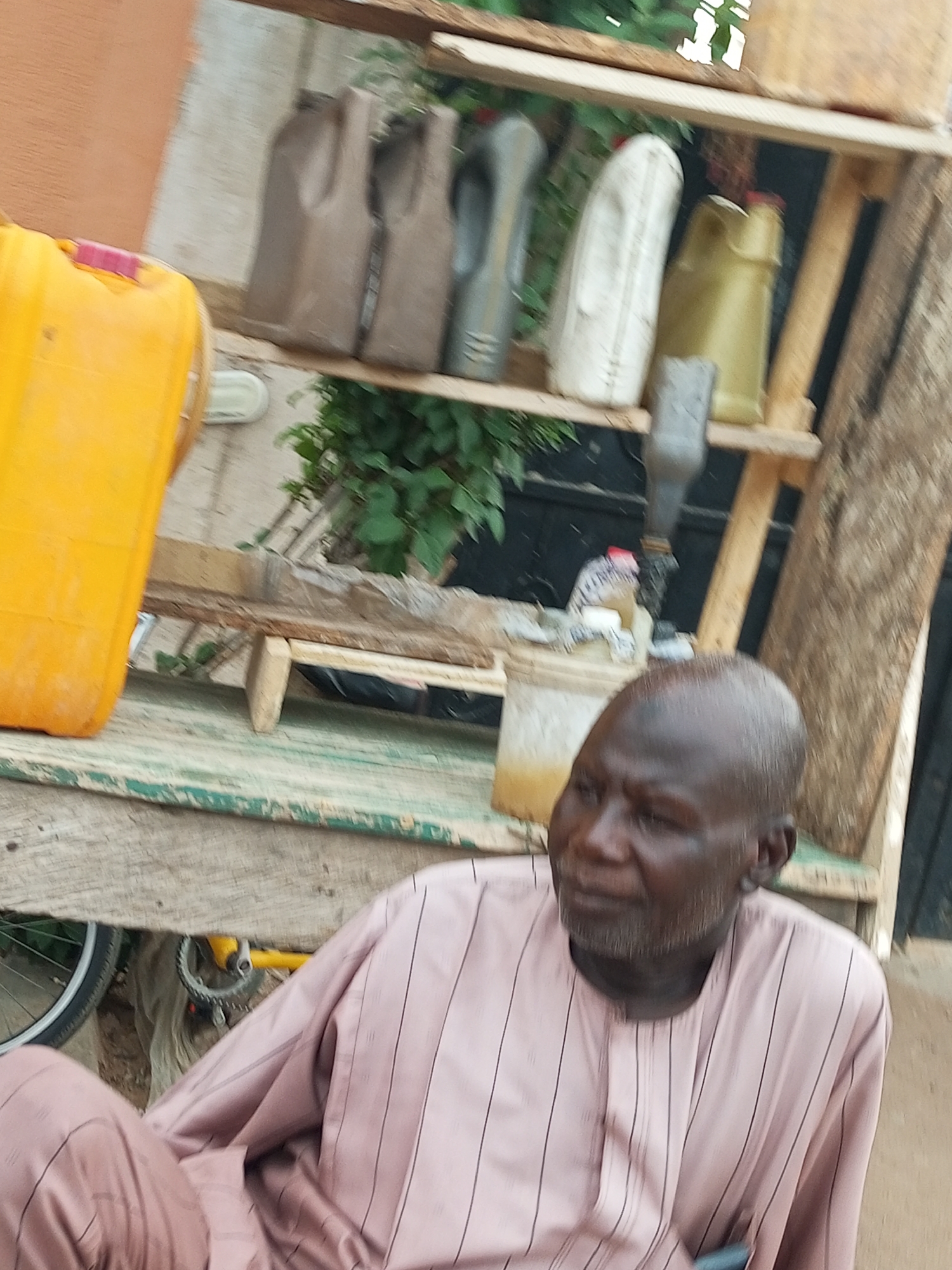 In Arawa, Nigeria, a man wearing a light pink, vertically striped tunic sits outdoors near a rustic wooden shelf unit. The shelf unit is stocked with various containers, including several jerrycans of different colors (yellow, green, grey, and brown) and a white, elongated bottle. In the foreground, a bicycle wheel is visible. The man is looking off to the right of the frame with a neutral expression. The environment appears to be informal, possibly a workshop or storage area, with visible greenery behind the shelves.