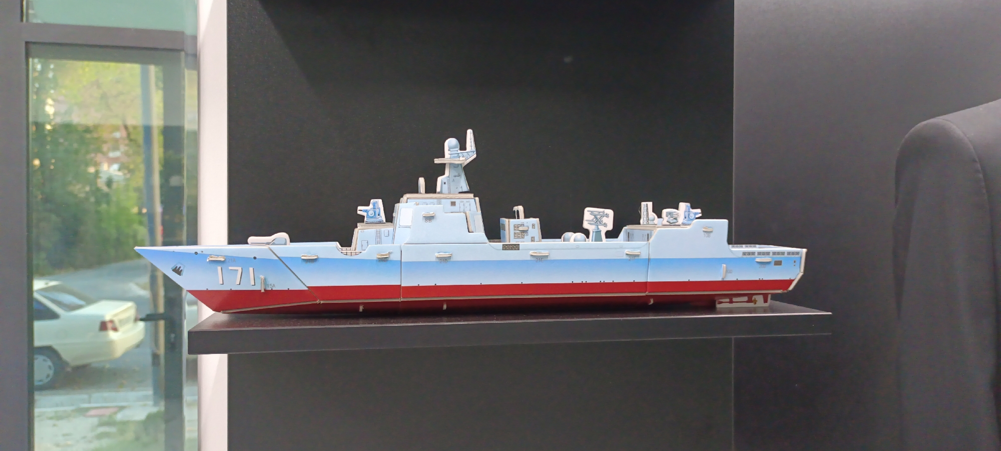 A light blue and red model of a warship sits on a black shelf against a dark gray background. The warship has "171" printed on its bow in white. The model is made of what appears to be cardboard or thick paper, with multiple pieces joined together. Several small structures and details, such as what look like cannons or radars, are attached to the deck and superstructure of the ship. A portion of a person in a black suit jacket is visible on the far right side of the image. To the left of the warship, a large window or glass partition reflects a street scene with trees, buildings, and a white car. The lighting suggests it is daytime, possibly late afternoon or early morning due to the soft light. The setting appears to be an indoor display area or showroom, possibly in Fergana City, Uzbekistan, given the reflective environment and the possibility of an exhibition. There is no discernible weather or specific emotional cues.