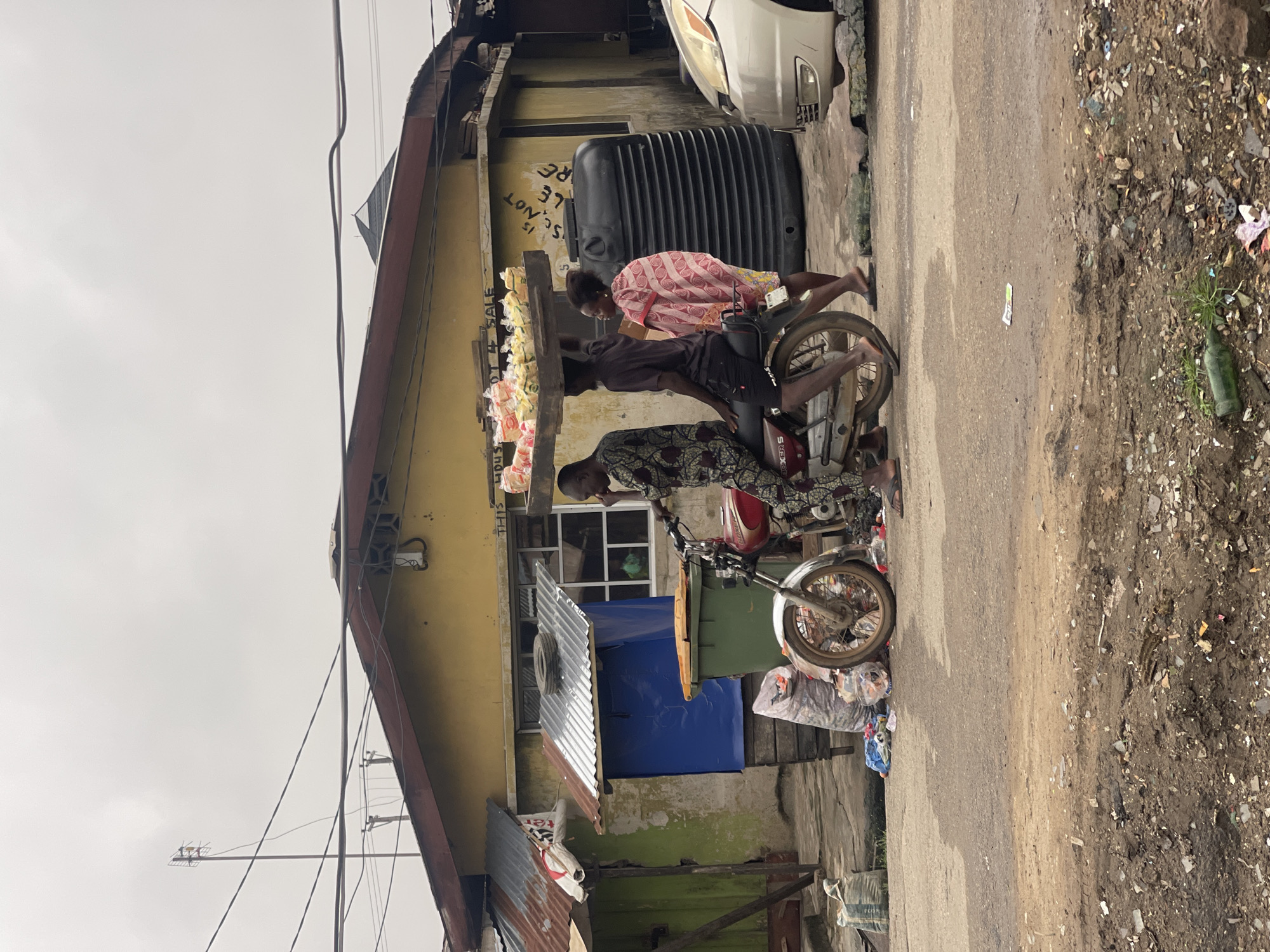 In Akowonjo, Nigeria, a street scene unfolds under an overcast sky. Three people are central to the image, two on a red motorcycle and one walking alongside. The motorcycle is a Boxer 150 model and is ridden by a man wearing a patterned dashiki. Another man, also in a dashiki, sits behind him, balancing a large wooden tray laden with numerous small, packaged items, likely food or goods, on his head. A woman in a pink and white patterned garment walks to the right of the motorcycle.
The environment is a dusty, unpaved street. In the background, a yellow building with a brown-tiled roof and corrugated metal extensions dominates the scene. Various structures and objects are scattered around, including a large black water tank to the right of the building, a green refuse bin, and piles of plastic bags and debris. A silver car is parked on the right side of the frame. Electrical wires crisscross above the structures. The ground is uneven and littered with assorted refuse, including plastic bottles and debris.