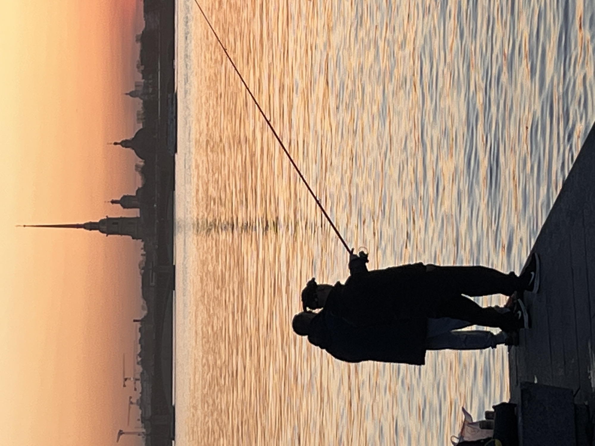 Two people are silhouetted fishing on a pier or embankment at sunset or sunrise. The silhouette of the Peter and Paul Fortress, a landmark in St. Petersburg, Russia, is visible across the water. The water reflects the colors of the sky. The pier is constructed of wooden planks. The person in the foreground is holding a fishing rod.