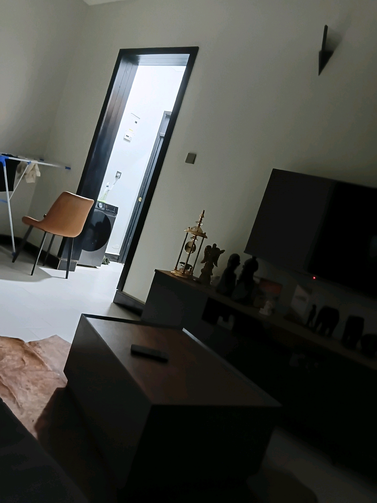 This image shows an interior scene, likely a living room and an adjacent laundry area, in Kampala, Uganda. In the foreground, a dark, angular coffee table sits on a patterned rug. To the left, a modern, orange-brown chair with black legs is positioned near a clothes drying rack. Beyond the chair, through an open doorway, a washing machine is visible in what appears to be a laundry room. The main living area features a dark entertainment unit displaying several decorative items, including small statues, and a large, dark television screen. A dark, arrowhead-shaped object is mounted on the light-colored wall above the entertainment unit. The lighting is dim, creating shadows throughout the room.