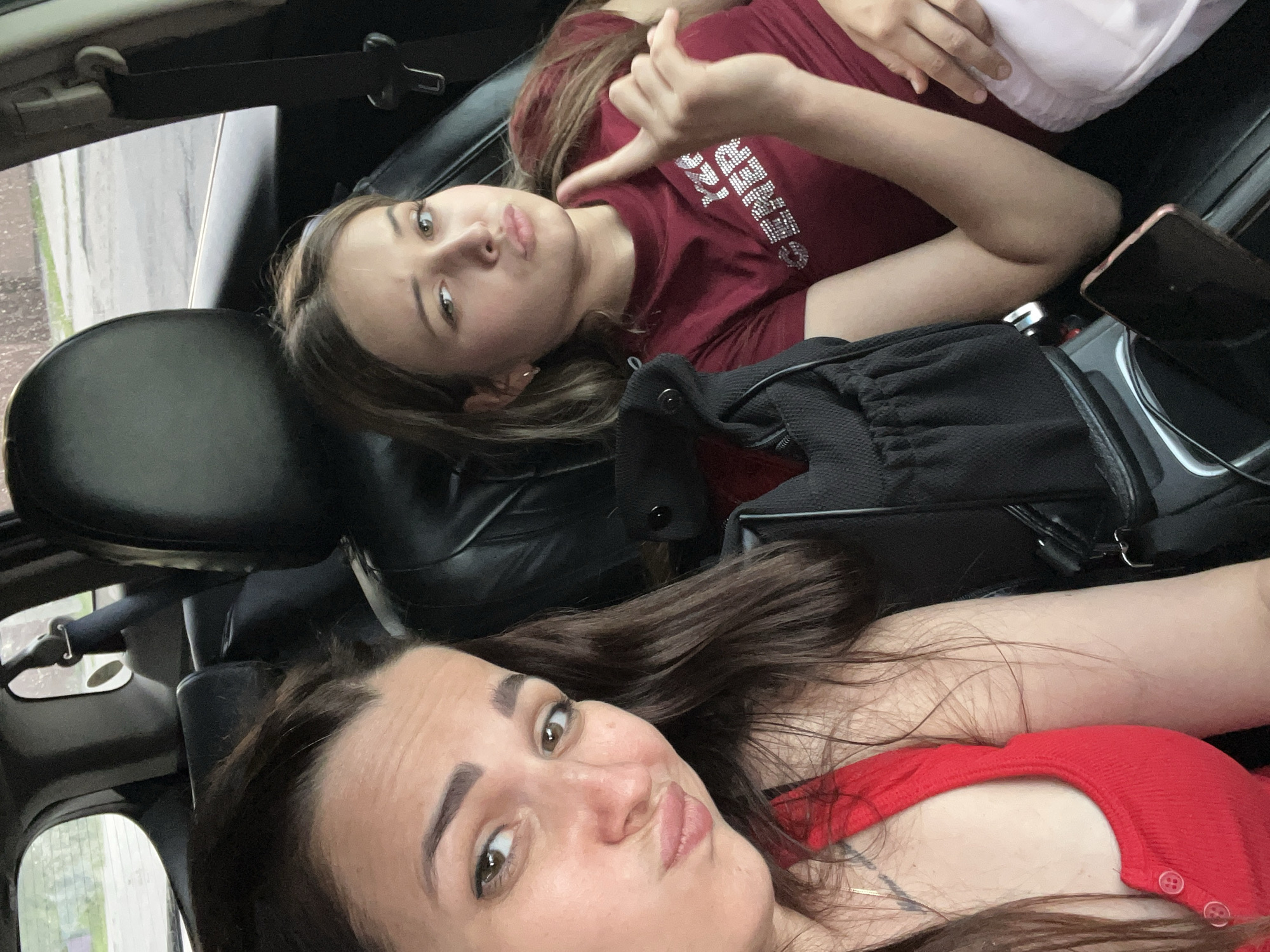 Two young women are inside a car, appearing to be in the front and back seats. The woman in the front seat, on the left, has long, dark brown hair and is wearing a red sleeveless top. She is looking at the camera with her lips pursed. The woman in the back seat, on the right, has long, light brown hair and is wearing a dark red t-shirt with a rhinestone design. She is looking slightly off-camera and has her right index finger raised. A black bag with a drawstring is visible between the two women. A smartphone lies face down on a console in the foreground. The interior of the car, including leather seats and a seatbelt, is visible. Outside the car windows, a street and some greenery can be seen, suggesting an urban or suburban environment.