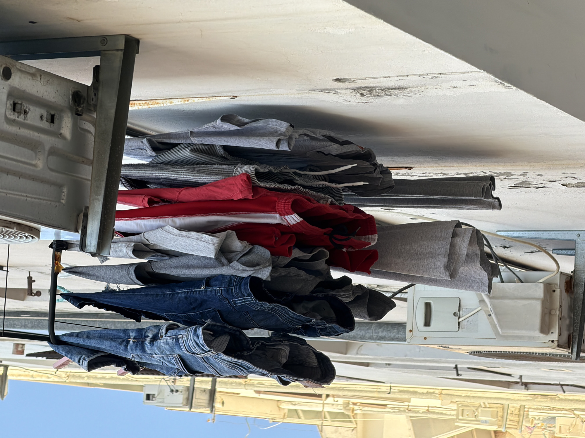 The image captures a close-up view of laundry hanging on a clothesline outdoors, in what appears to be a dense urban environment, possibly in Budva, Montenegro. A variety of clothing items, including denim jeans, t-shirts, and shorts, are visible, some fastened with pink and yellow clothespins. The clothesline is supported by a metal rod which is attached to the side of a building. In the background, elements of the building's exterior are visible, including what seems to be an air conditioning unit mounted on the wall and some electrical conduits. The lighting suggests it is daytime, with shadows cast on the textured, weathered wall. The overall scene depicts a common urban sight of drying laundry.