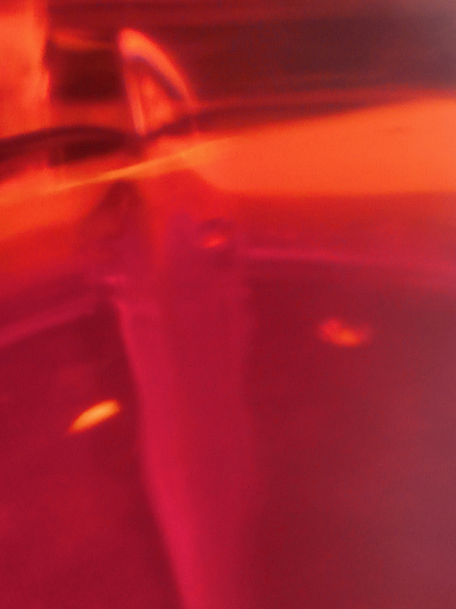 The image is an abstract, out-of-focus photograph dominated by shades of red and orange, suggesting a play of light and shadow on a reflective surface. There are no discernible landmarks or features that would place it within any specific city, including Delhi, India. The composition is devoid of people, objects, or discernible actions, rendering a forensic description impossible.
