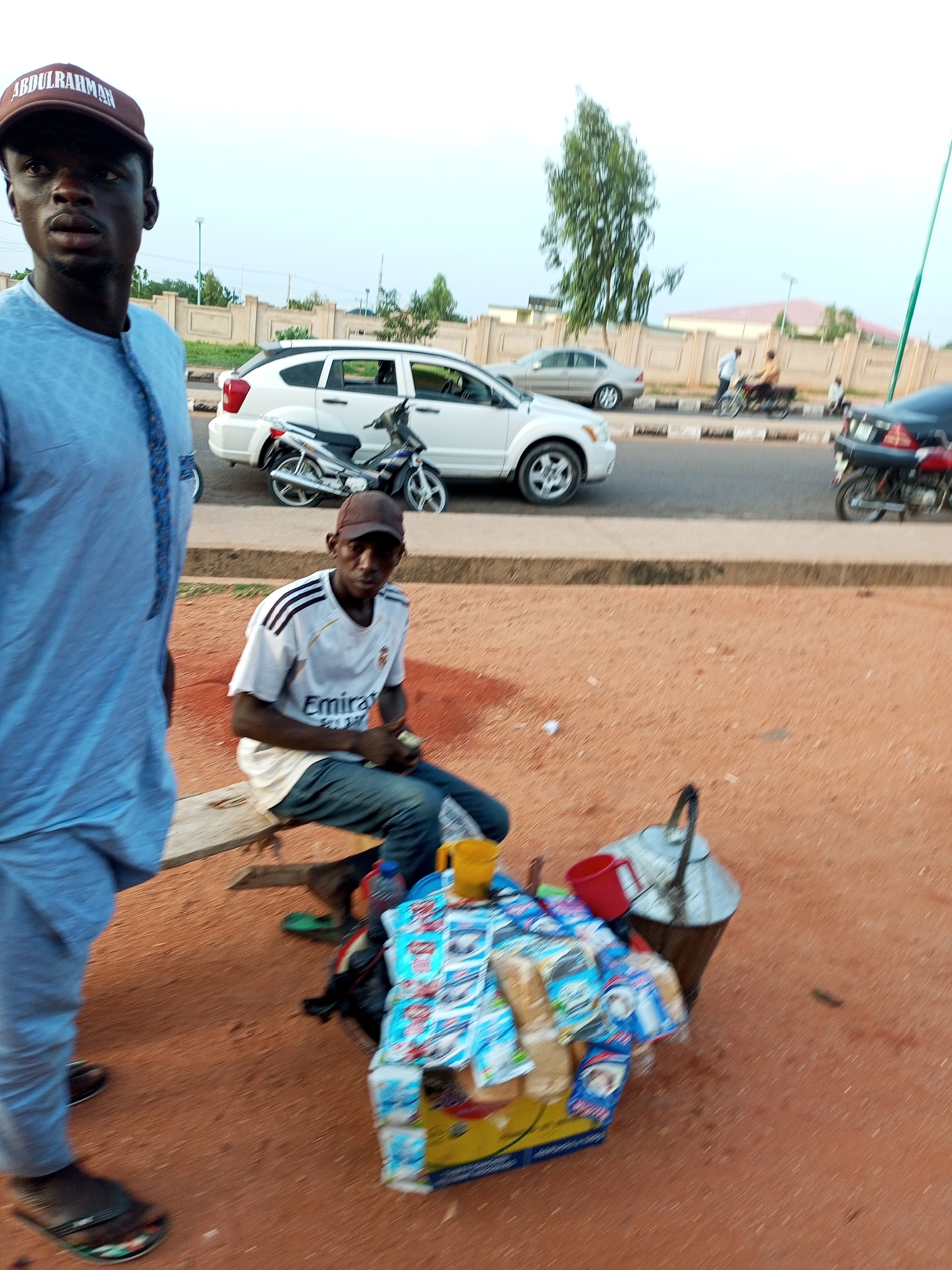 This media file features two individuals in an outdoor setting, likely a roadside in a warm climate. The foreground is dominated by reddish-brown earth, suggesting a dry environment.
On the left, a person stands facing the camera, partially cropped out of the frame. They are wearing a light blue, long-sleeved tunic and a dark baseball cap with the word "ABDULRAHMAN" embroidered in white lettering. Their expression is neutral.
In the mid-ground, a second person is seated on a wooden bench. This individual is wearing a white t-shirt with black stripes on the sleeves and the word "Emirates" printed across the chest. They are looking towards the camera with a calm demeanor, holding a few small items in their hands.
In front of the seated person, there is a collection of items that appear to be goods for sale. These include several packaged food items with colorful labels, a yellow cup, a red container, and a large metal kettle. A water bottle is also visible.
Behind the individuals, a road is visible with several vehicles. A white SUV is parked, and a motorcycle is in front of it. Further down the road, other cars and a motorcycle are moving. The background shows a wall, trees, and buildings, indicating a built-up area.
The lighting suggests it is daytime, likely late afternoon given the soft light. The weather appears to be clear. The overall scene depicts a moment of everyday life, possibly a vendor waiting for customers or taking a break.