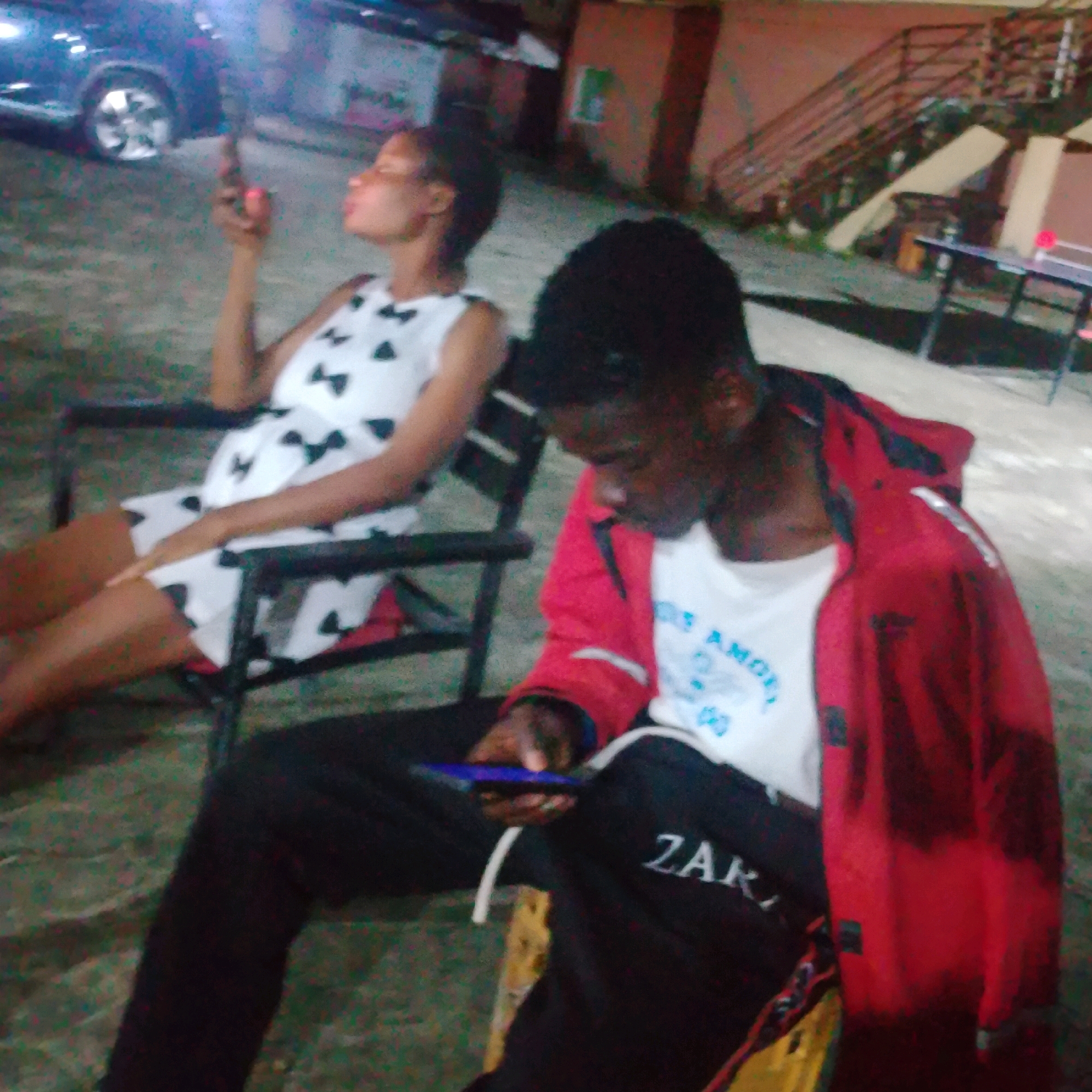 In Ado, Nigeria, two individuals are seated outdoors in the evening. The person on the left, appearing to be female, is wearing a white dress with black bow patterns and is looking upwards while holding a mobile phone. To the right, a person, likely male, is wearing a red jacket over a white t-shirt and black pants with "ZAK" printed on them, and is also looking at a mobile phone in their hand. They are both seated in chairs, with the chairs positioned close together. The environment suggests a casual outdoor setting, possibly a compound or a common area, with concrete flooring and hints of vegetation. A dark-colored vehicle is partially visible in the background on the left. The lighting indicates it is nighttime, with some artificial illumination present.